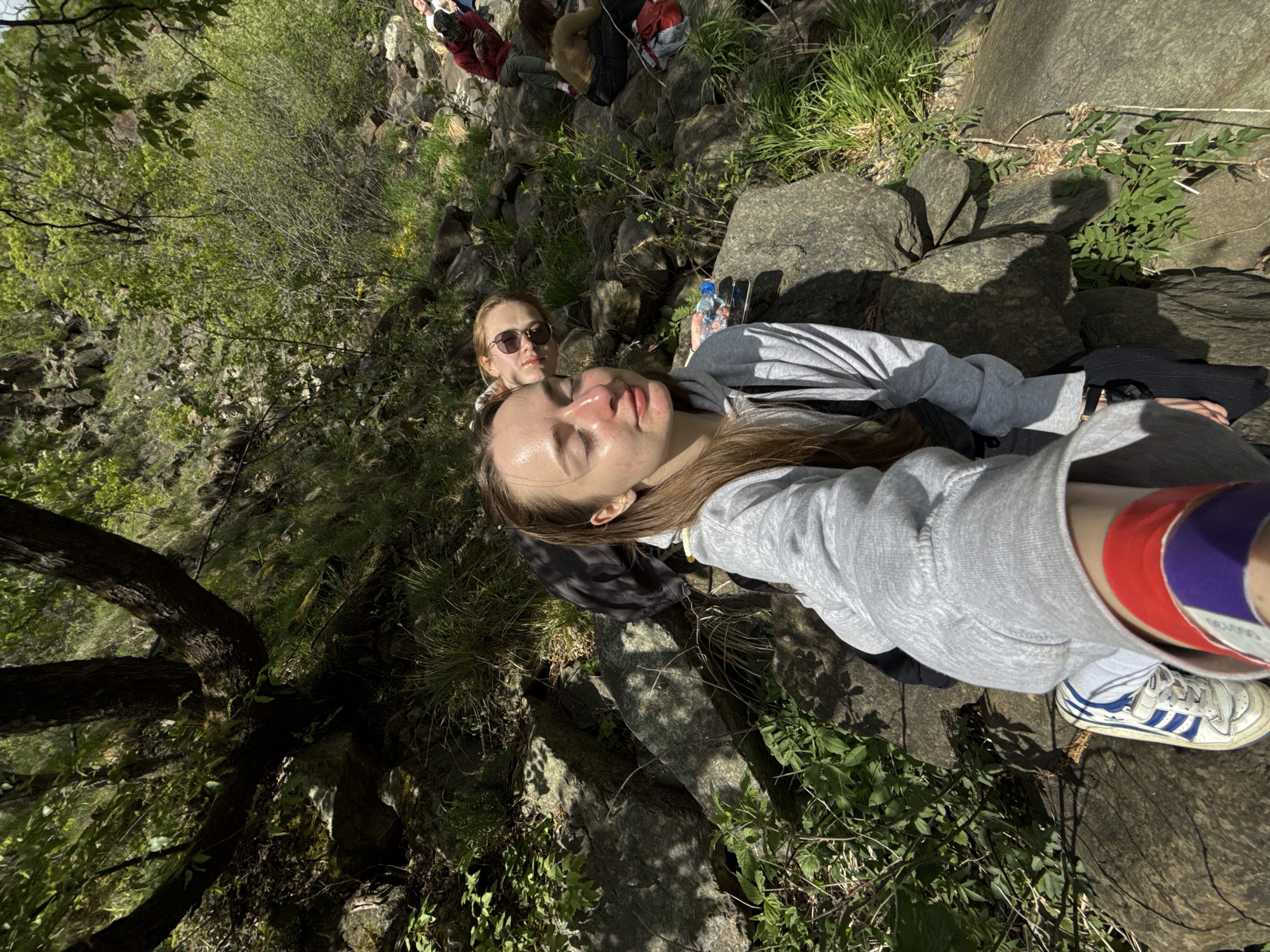 A tilted eye-level shot shows two women in an outdoor setting in Zamčanje, Serbia. One woman with closed eyes is in the foreground wearing a grey hooded sweatshirt with red and purple wristbands, and white Adidas sneakers with blue stripes. Behind her is another woman wearing sunglasses, with a water bottle next to her. Both are positioned amongst rocks, grass, and sparse foliage. In the background, out of focus, there are several people sitting or lying down. A tree is visible on the left side of the image.