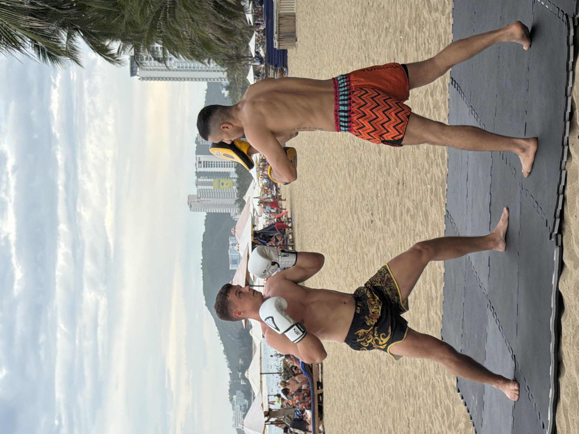 Two shirtless men are sparring on a black mat on a sandy beach in Nha Trang, Vietnam. The man on the left is wearing black Muay Thai shorts with gold trim and white boxing gloves. He is in a defensive stance, looking to his right. The man on the right is wearing orange and black chevron-patterned Muay Thai shorts. He is holding a yellow and black punch pad in his left hand, ready to strike. In the background, there are beach umbrellas, lounge chairs, people relaxing on the sand, and several tall buildings. Palm trees are visible to the right of the frame. The sky is overcast with scattered clouds.