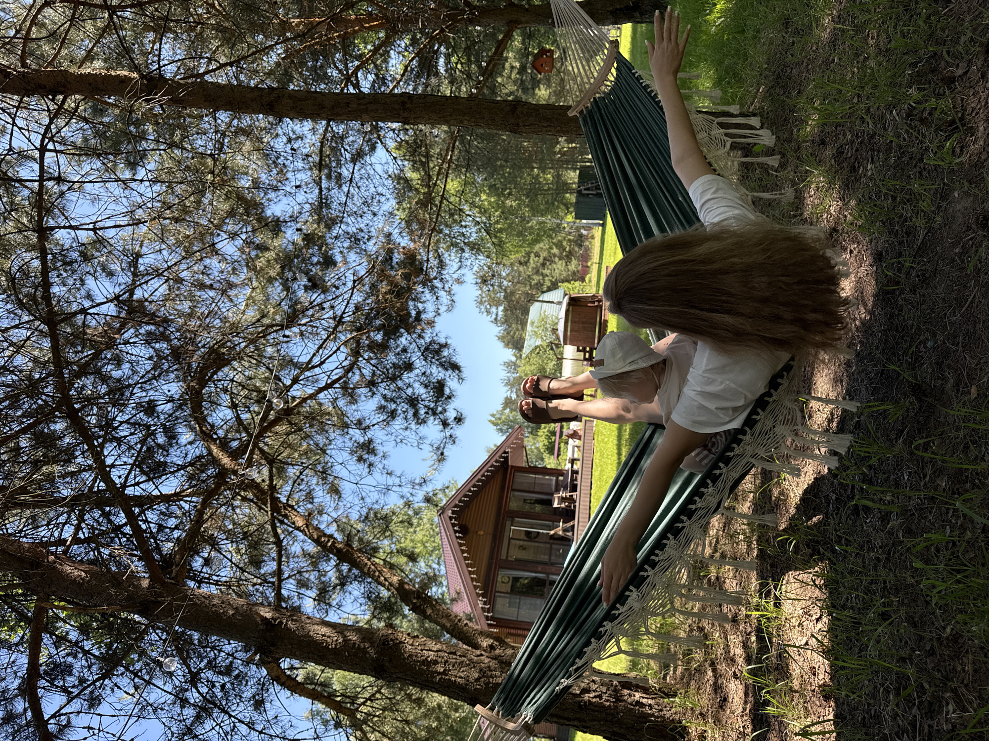 The scene depicts an outdoor environment in Копнино, Russia, on a clear, sunny day. One individual is observed lying on their back in a green fabric hammock with white braided rope and tassels, suspended between two large pine trees. The person's head is oriented towards the left, and their feet towards the right. They possess long, light brown hair extending down their back and are wearing a white short-sleeved top and dark brown open-toed sandals or flip-flops. A light-colored cap is visible on their head. Their right arm is extended outwards, resting on the hammock's edge, with fingers slightly curled.
The ground beneath the hammock consists of sparse green grass interspersed with brown earth and pine needles. Bright sunlight filters through the tree canopy, creating distinct shadows on the ground. The sky overhead is clear and blue, visible through the branches of multiple pine trees.
In the mid-ground, a two-story wooden house with a dark brown pitched roof is visible. This structure features multiple large windows, a glass door, and a porch or deck area with outdoor furniture. Ornate white trim lines the eaves of the roof. Further to the right and slightly behind, a smaller, rustic wooden structure with a lighter, possibly green, domed roof is present. In the far background, behind the secondary structure, a green shed or container is visible.
A string of clear-bulbed decorative lights is strung between the trees in the upper right portion of the image. A wooden birdhouse is also suspended from a tree branch in this area. Lush green foliage and distant trees complete the horizon.