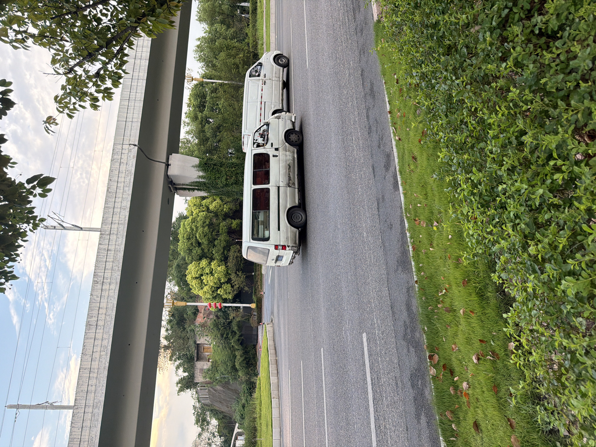The image depicts a street scene in Jiangjin District, Chongqing, China. Two white vans are traveling on a multi-lane asphalt road. The van on the left is older and appears to have some damage or wear on its side. The van on the right is also white and in better condition. Overhead, a large concrete overpass or bridge spans the road, with power lines running alongside it. Green trees and vegetation line the sides of the road and cover a hillside in the background. The sky is partly cloudy, suggesting it could be late afternoon or early morning. There are some fallen leaves scattered on the grassy median and the roadside. In the distance, a small, weathered building with a yellow door is visible on the hillside, with a red traffic signal pole nearby. The overall impression is of a moderately busy urban or suburban road with some natural elements present.