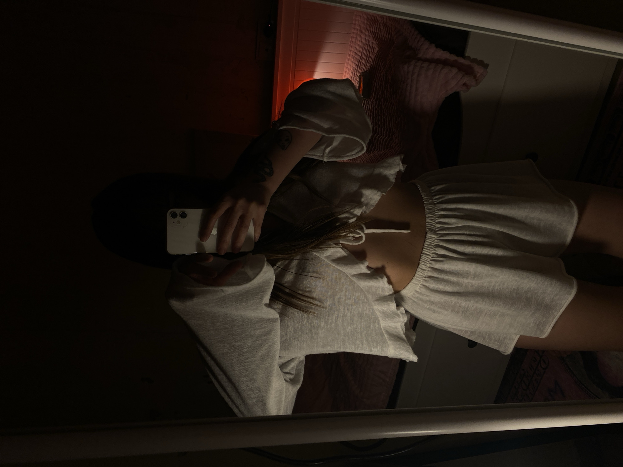 A woman is reflected in a mirror, holding a white smartphone in her left hand. She is wearing a light grey, short-sleeved, ruffled top and matching shorts. Her dark hair is partially visible. The reflection shows a portion of a room with a pink, textured blanket or pillow behind her. The overall lighting is dim with a distinct contrast. The location cannot be determined as Votkinsk, Russia as the location is not visible in the image.