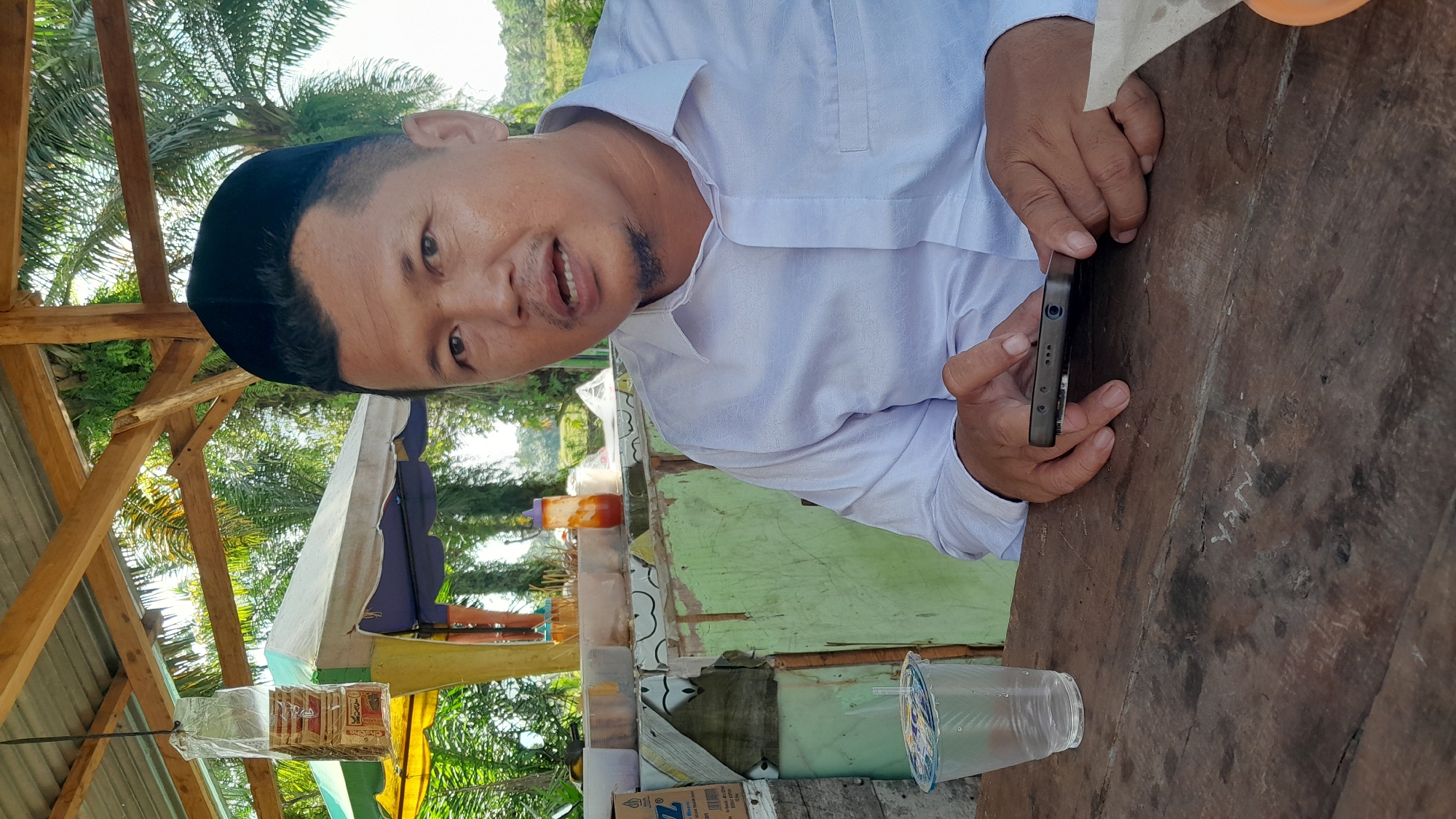 The image shows a man wearing a black peci hat and a white shirt, holding a black smartphone in his hands. He is standing behind a wooden table. In front of him, there is a small stall or counter with various items displayed, including bottled drinks, packaged goods, and a plastic cup. The background includes trees and a wooden structure, suggesting an outdoor setting. The location is the city of Blangriek, Indonesia.