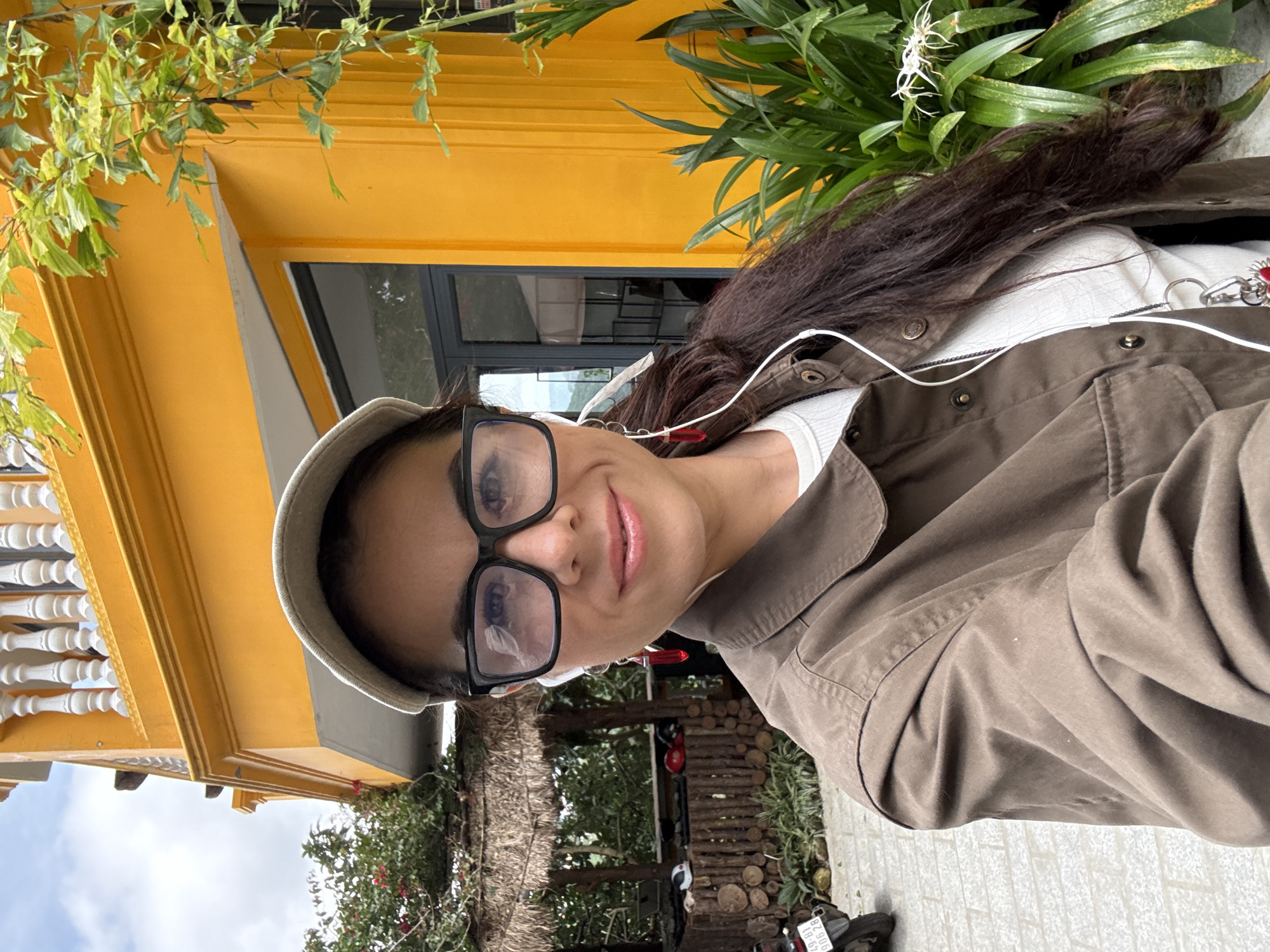 In Da Lat, Vietnam, a woman with long dark hair, wearing a brown jacket, a white top, a beige baseball cap, and large black-rimmed glasses, is taking a selfie outdoors. She has white earbuds with red dangling earrings. The background features a brightly colored yellow building with white balustrades on an upper level and lush green foliage. To the left, there is a rustic structure made of wood and thatch, possibly a shelter or kiosk, with a motorcycle parked nearby. The ground appears to be paved with stones.
