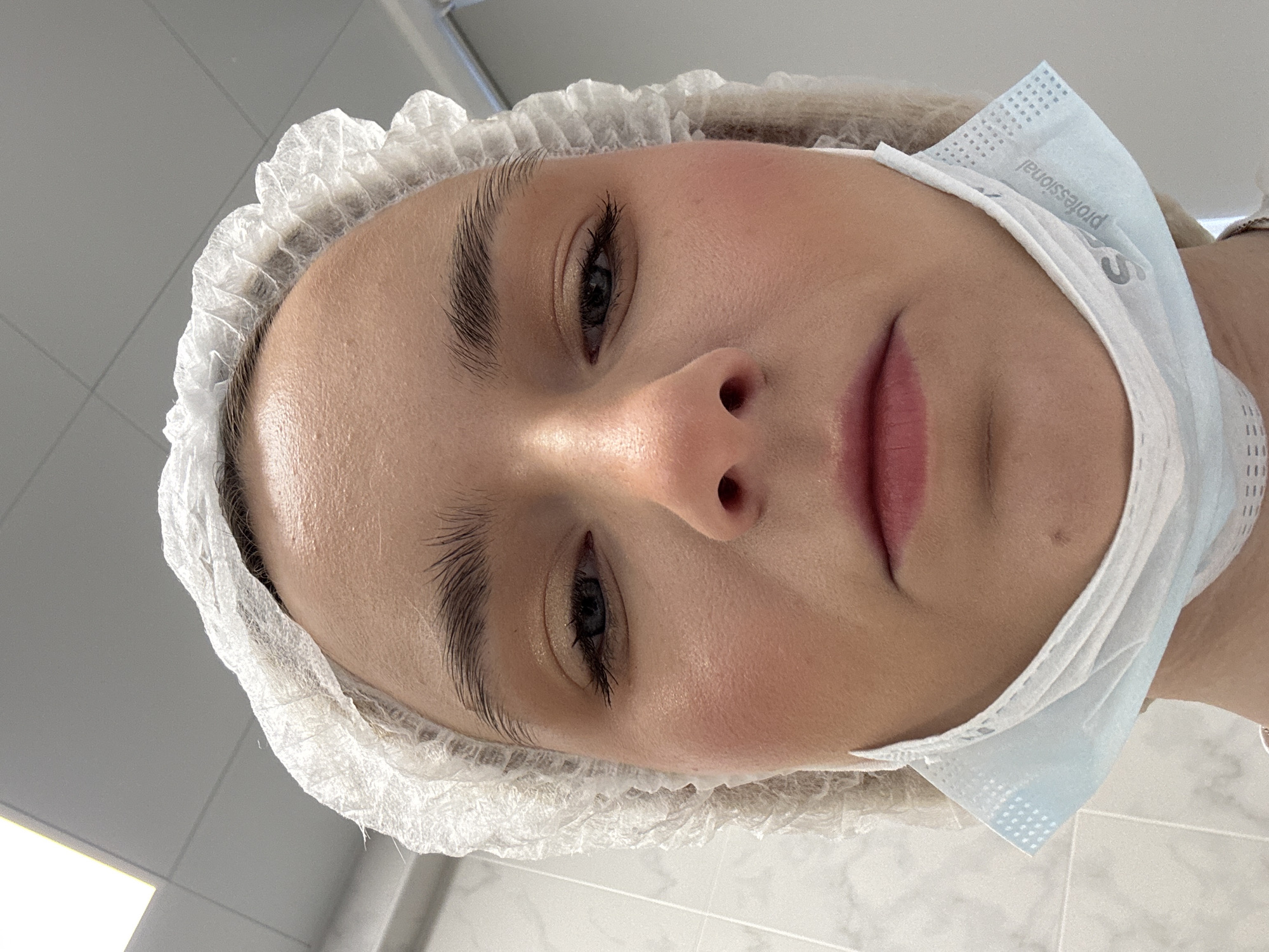 A person wearing a white surgical cap and a light blue surgical mask that has been pulled down under their chin is visible in a close-up photograph. The person has light skin, dark eyebrows, and eyelashes. The lips are a soft pink color. The background appears to be a white or light-colored tiled wall. The mask has the word "professional" printed on it.