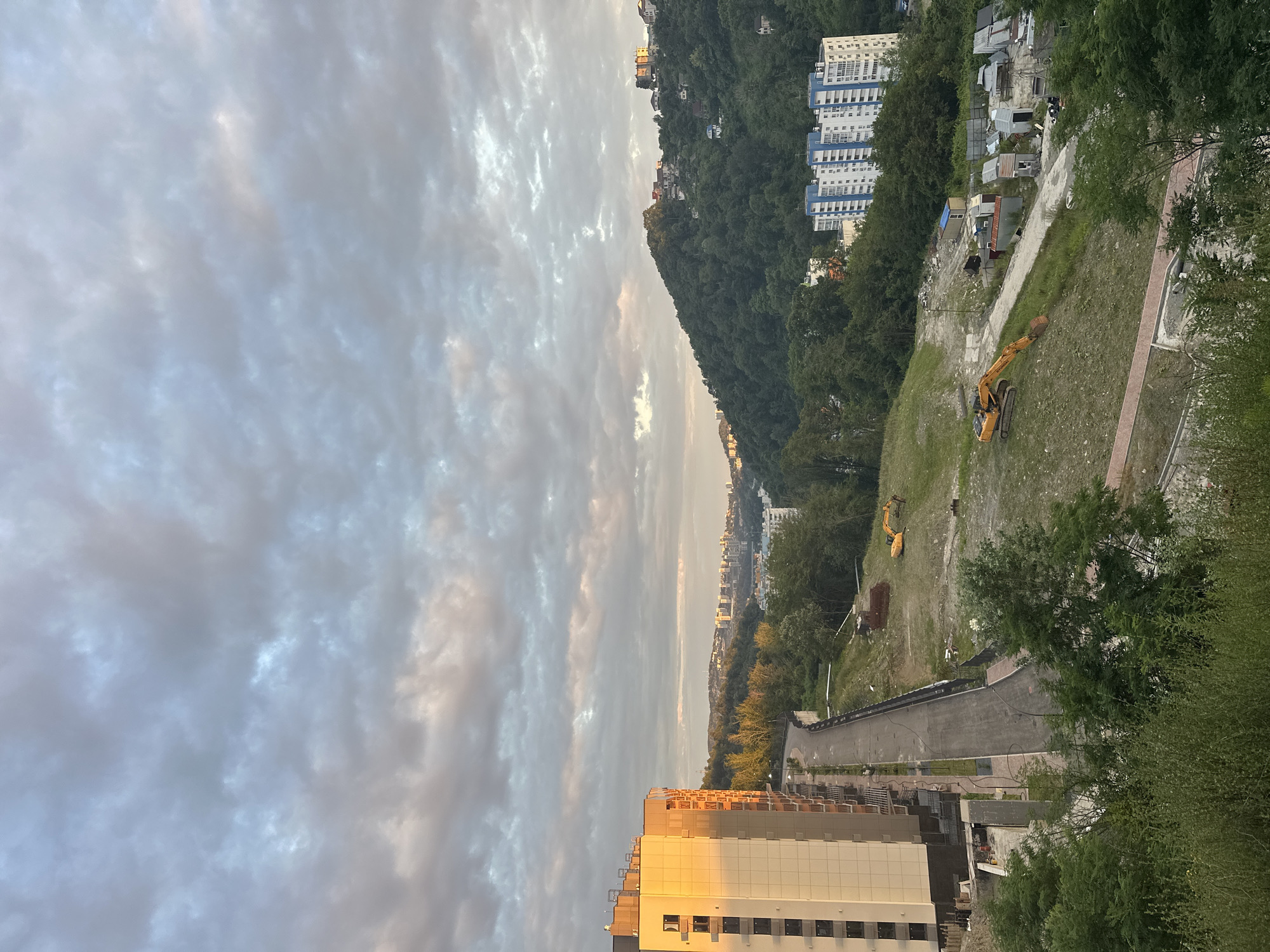 The image captures a panoramic view of Razdolnoye, Russia, during what appears to be late afternoon or early evening. The scene is dominated by a developing urban landscape situated amidst rolling green hills.
In the foreground to the left, a modern, multi-story building under construction is prominent. Its facade is a mix of white panels and darker accents, with balconies visible. Sunlight casts a warm golden glow on the side of this building, indicating the direction of the setting sun. Adjacent to this building is a paved road that winds uphill.
The middle ground of the image is characterized by a construction site. An excavator, painted bright yellow, sits on a grassy slope. Scattered debris and temporary structures, possibly associated with the construction, are visible in this area. More buildings, some with blue and white facades, are situated further back, nestled among the trees.
The background features a lush, densely forested hillside dotted with houses and buildings, suggesting a residential area that climbs the slopes. A distant city skyline is faintly visible on the horizon. The sky is filled with a dramatic display of clouds, painted in shades of gray, white, and hints of pink and orange, characteristic of a sunset. The overall impression is one of a city undergoing development, set against a backdrop of natural beauty.