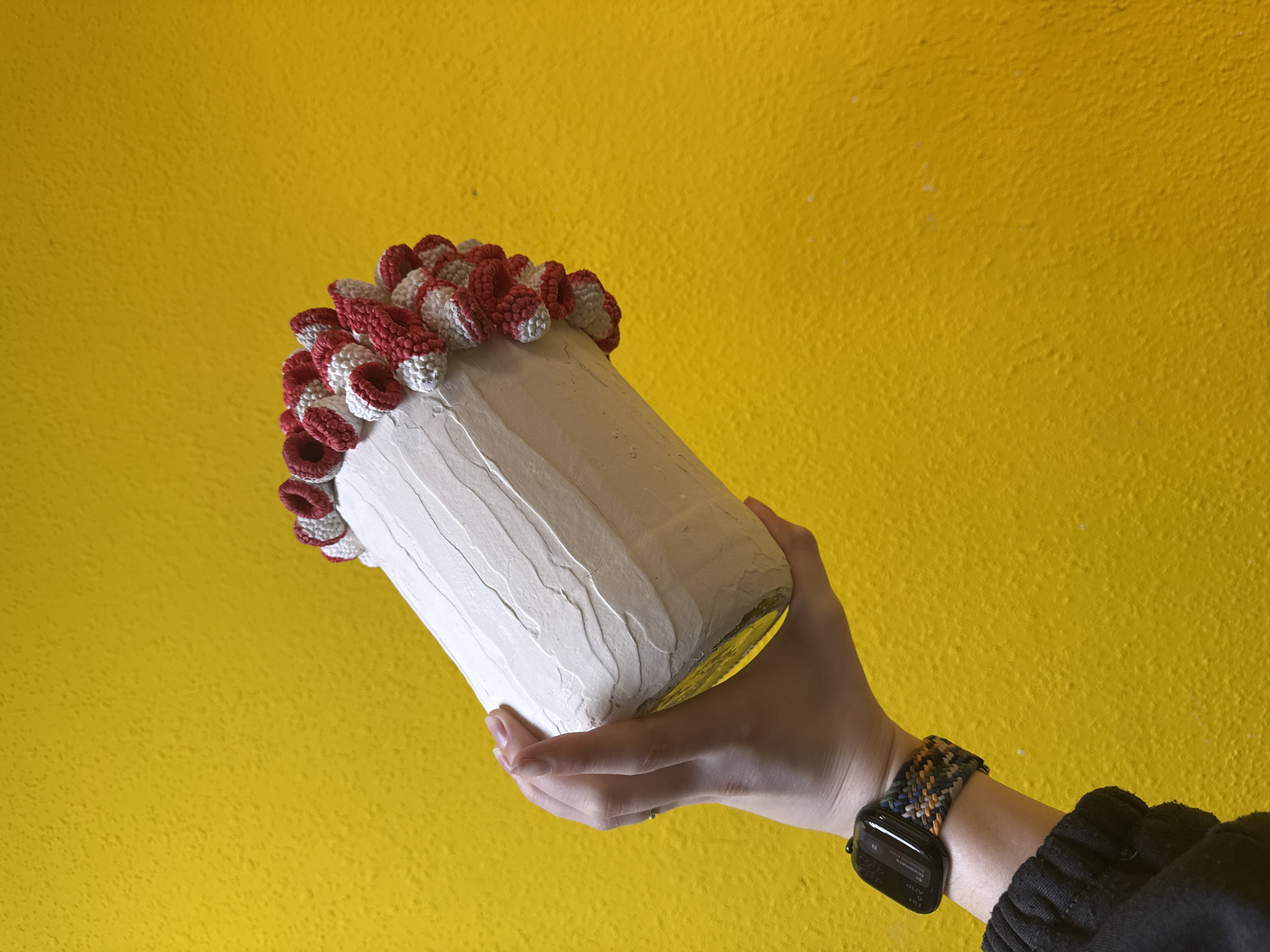A hand holding a decorated glass jar is visible against a textured yellow background. The jar is cylindrical and coated with a white, textured material, possibly plaster or paint. The top rim of the jar is adorned with crocheted elements in red and white. The hand holding the jar has an arm extending from the bottom right of the frame, wearing a smart watch with a woven band. The scene appears to be indoors, given the wall as a backdrop, but the specific location or city is not discernible from the image.