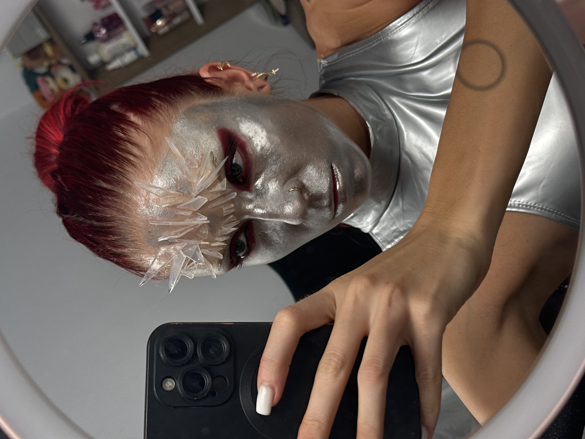 The image is a close-up selfie taken by a person with bright red hair styled in a bun. The person has applied silver body paint to their face and neck, with bold red eyeshadow and metallic silver lipstick. A cluster of clear, sharp-edged acrylic shards is attached to their forehead and temples, resembling ice. They are wearing a shiny, metallic silver, sleeveless top. The person is holding a black smartphone in their left hand, capturing the reflection in a ring light.
The background shows a slightly out-of-focus shelf filled with various bottles and containers, suggesting a personal vanity or a beauty setup. The environment is an indoor setting, likely a room. There is no indication or visual evidence to suggest this scene is located in North Kuta, Indonesia.