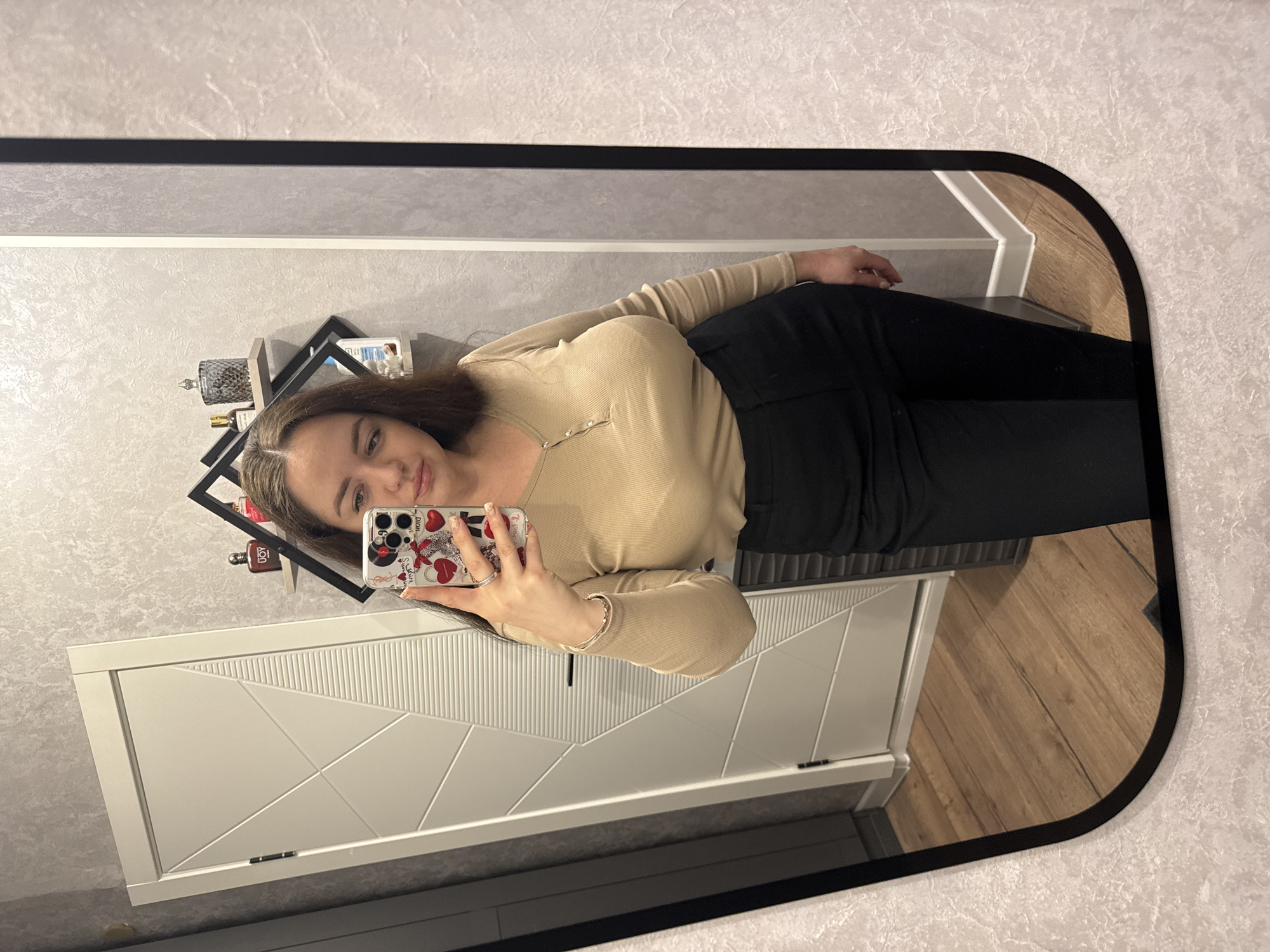 A young woman is taking a selfie in a mirror. She is wearing a beige long-sleeved top and black pants. She is holding her phone, which has a heart-patterned case. Behind her, there is a white cabinet with a geometric design and a black shelf with various items on it. The wall is decorated with a light-colored patterned wallpaper. The floor is made of wooden planks. The location is Yakhroma, Russia.