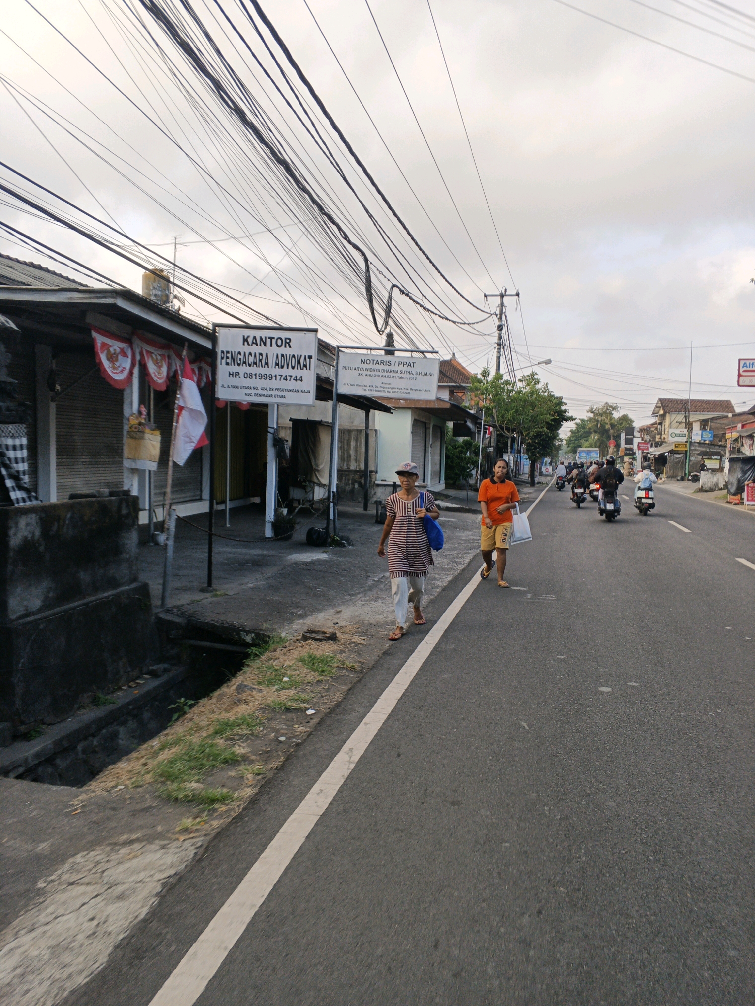 This is a street-level photograph captured in Denpasar, Indonesia. The scene is dominated by a road with a white line marking the edge, bordered by a sidewalk and buildings on the left. Overhead, a dense network of electrical wires crisscrosses the sky.
On the sidewalk, two women are walking. The woman closer to the foreground, on the left, is wearing a striped dress and a cap, and carries a blue bag. The woman to her right wears an orange t-shirt and yellow shorts and carries a white bag. Several motorcycles are traveling on the road in the background.
The buildings on the left appear to be commercial establishments. One building has a sign that reads "KANTOR PENGACARA/ADVOCAT" and another sign for "NOTARIS/PPAT". Flags with the Indonesian red and white colors are visible. The environment suggests a typical urban street scene in Indonesia, with a mix of businesses, traffic, and pedestrian activity. The time of day appears to be either early morning or late afternoon, given the soft lighting and the overcast sky.