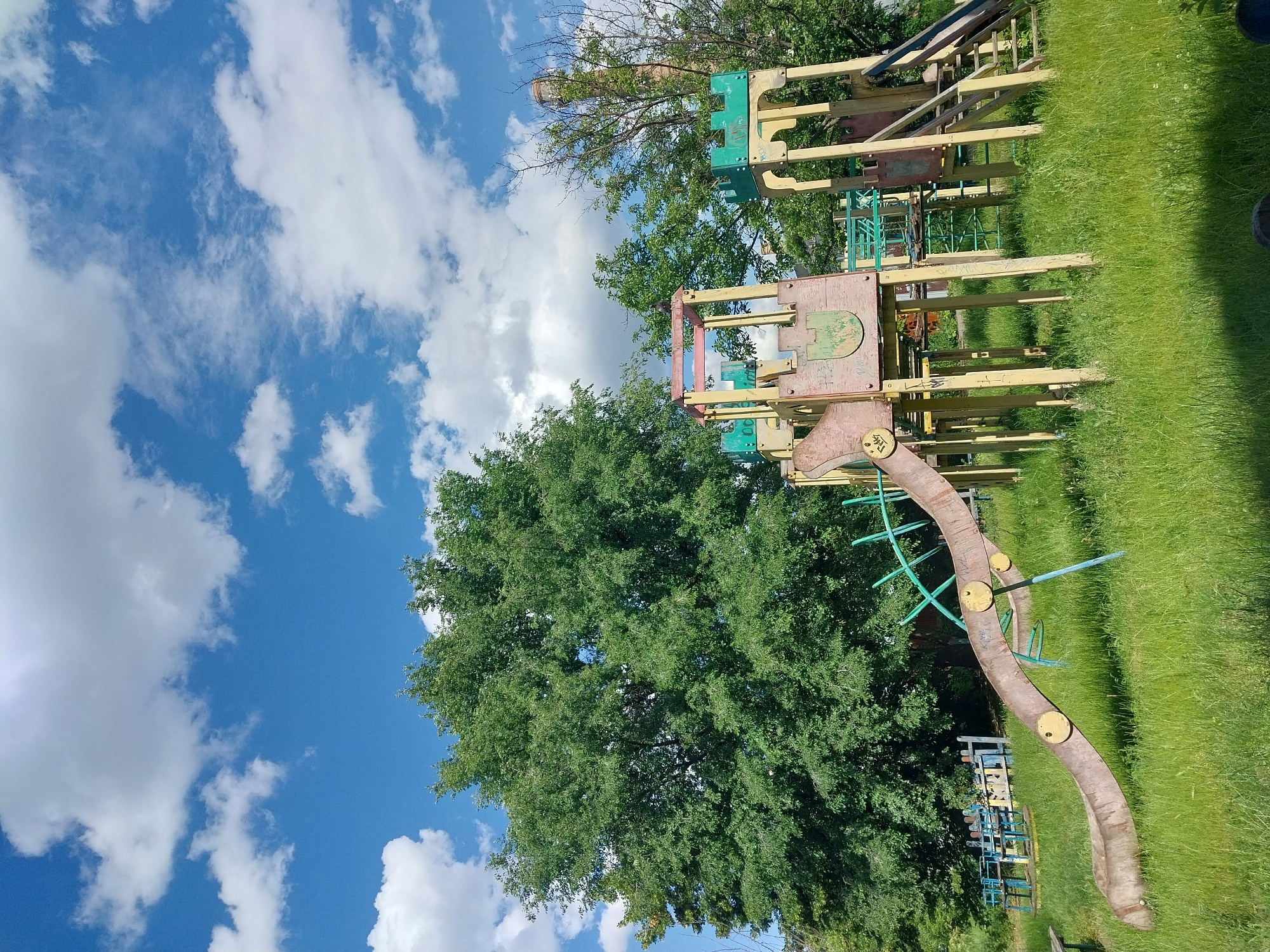 A weathered, multi-colored playground structure, resembling a castle, stands in a grassy field under a bright blue sky dotted with fluffy white clouds. The structure is made of wood and metal, with a long, curved slide extending from the left side of the frame towards the foreground. Yellow and green paint adorn parts of the structure, some of which is peeling. To the left of the main structure, a set of blue and yellow swings is visible. A large, leafy green tree dominates the background on the left, while other trees and a tall brick chimney are visible on the right. The grass in the field is long and somewhat unkempt. The scene appears to be in an outdoor public space, likely a park or playground in Dnipro, Ukraine, given the context. There are no people or animals visible in the image.