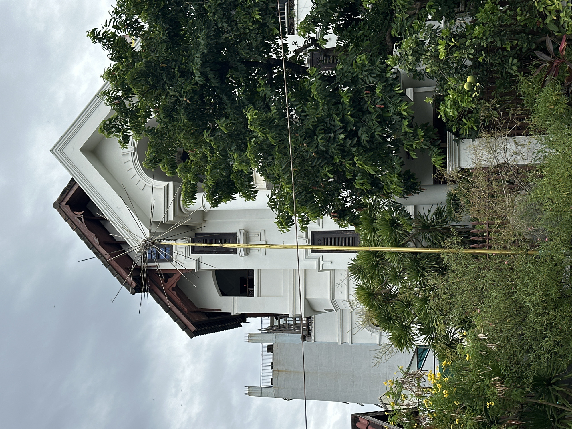 The image depicts a white building with traditional architectural elements in Da Nang, Vietnam. The building features a steeply pitched wooden roof, decorative cornices, arched windows, and shutters. A prominent gold-colored pole with a starburst-shaped finial stands in front of the building. Lush green trees and foliage surround the structure, with some yellow flowers visible on the left. In the background, a tall, lighter-colored structure with a railing on top is partially visible. The sky is overcast. The scene appears to be a residential or commercial property, possibly a hotel or guesthouse, given its aesthetic. The overall impression is of a well-maintained property with a tropical ambiance.
