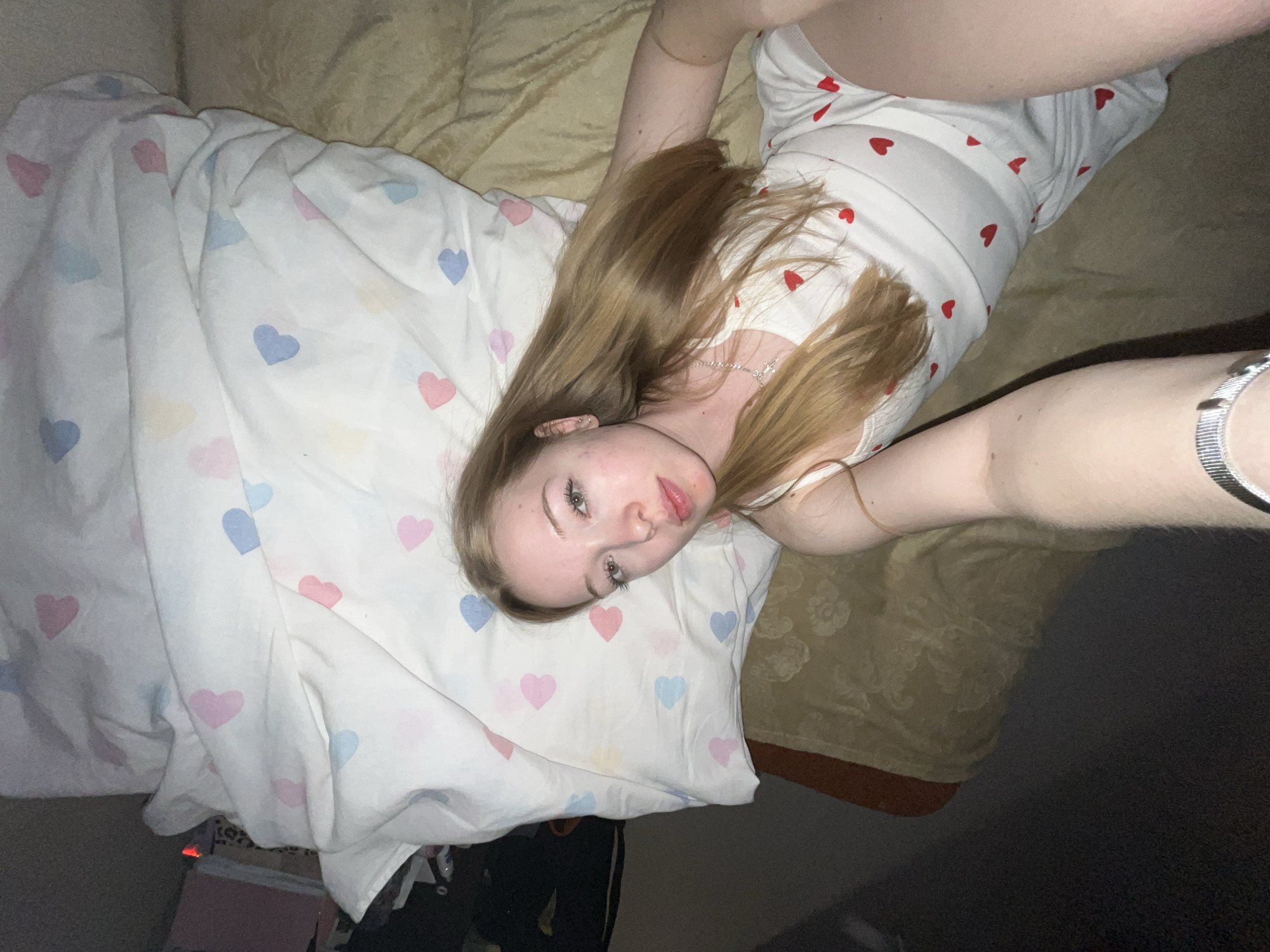 A young woman is lying on a bed in what appears to be a bedroom in Chaikovsky, Russia. She has long, light brown hair and fair skin, looking directly at the camera with a neutral expression. She is wearing a white top adorned with small red heart patterns. Her left arm is extended, holding what seems to be the camera for a selfie, revealing a silver watch on her wrist. Around her neck is a delicate silver necklace with a small charm, possibly a cross.
She is partially covered by a white duvet or blanket featuring a cheerful pattern of blue, pink, and yellow hearts. Beneath her, a beige or gold-colored patterned fabric, likely another blanket or bedspread, is visible. The lighting in the room appears dim, suggesting evening or low light conditions. Some indistinct objects are partially visible on the floor next to the bed. The overall scene conveys a relaxed and personal atmosphere. There is no visible text in the image.