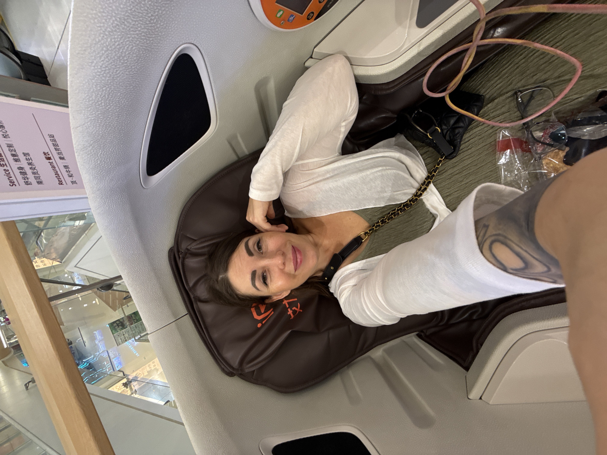 A woman is taking a selfie in a massage chair within a commercial establishment. She is seated in a reclining position, facing the camera and smiling. She is wearing a green dress and a white cardigan. A black quilted purse with a gold chain strap is visible on her right side. A pair of glasses and a bag of snacks are on the floor of the chair. The massage chair appears to be in a public area, possibly a mall or an airport, with signage in Chinese in the background. The overall environment suggests a commercial setting with modern amenities. The specific location in Huizhou, China is not explicitly identifiable from the image content.