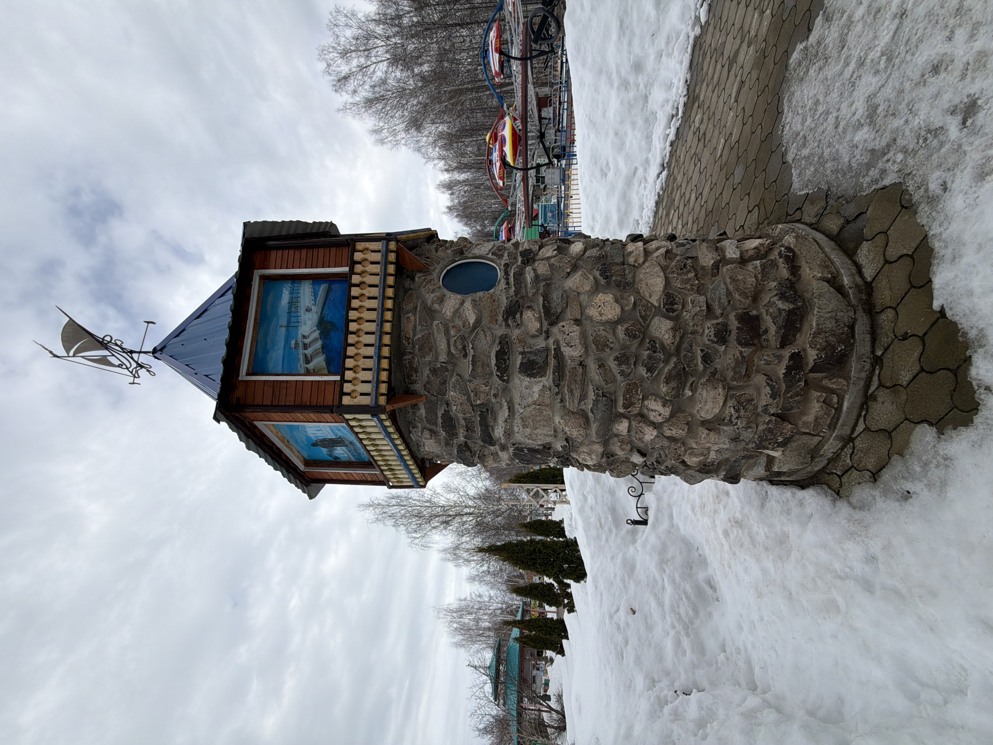 The image shows a decorative tower in the city of Chaikovsky, Russia. The tower has a stone base and a wooden top section with framed pictures. A weather vane sits atop the tower. Snow is present on the ground. In the background, a carousel can be seen. The sky is overcast. A paved path runs alongside the tower. There are several small evergreen trees.