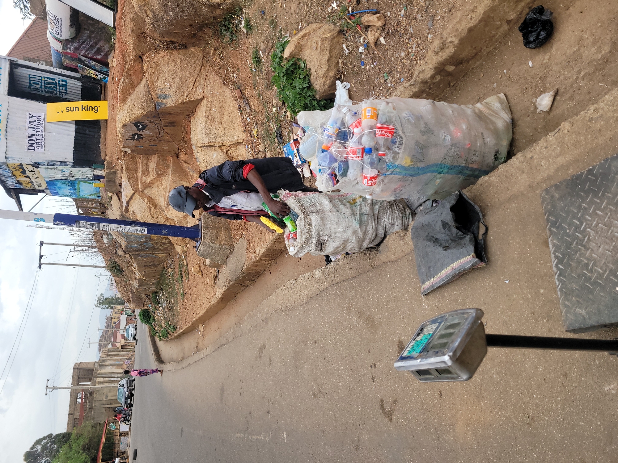 The image depicts a roadside scene in Jos, Nigeria, where a man is engaged in sorting plastic bottles. The man, wearing a dark jacket, red shirt, and grey cap, stands amidst large, transparent bags overflowing with collected plastic waste, including numerous "Coca-Cola" branded bottles. He appears to be actively handling the bottles, likely for recycling or resale. A digital weighing scale with a metal platform is visible to the right, suggesting the collected items are being weighed.
The setting includes an asphalt road in the foreground and a raised dirt embankment on the left, which is strewn with various debris and sparse green vegetation. Further along the embankment, a makeshift structure is visible, featuring signs. One prominent sign reads "DON JAY ART STUDIO" along with phone numbers: "08107451881, 08129385258". Another bright orange sign displays the text "sun king". In the distance, the road curves, with a person in a pink and white patterned outfit walking. Buildings, some appearing under construction, and utility poles with power lines line the distant roadside. The sky is overcast, indicating it is daytime. The overall scene reflects an informal economic activity related to waste collection in a semi-urban environment.