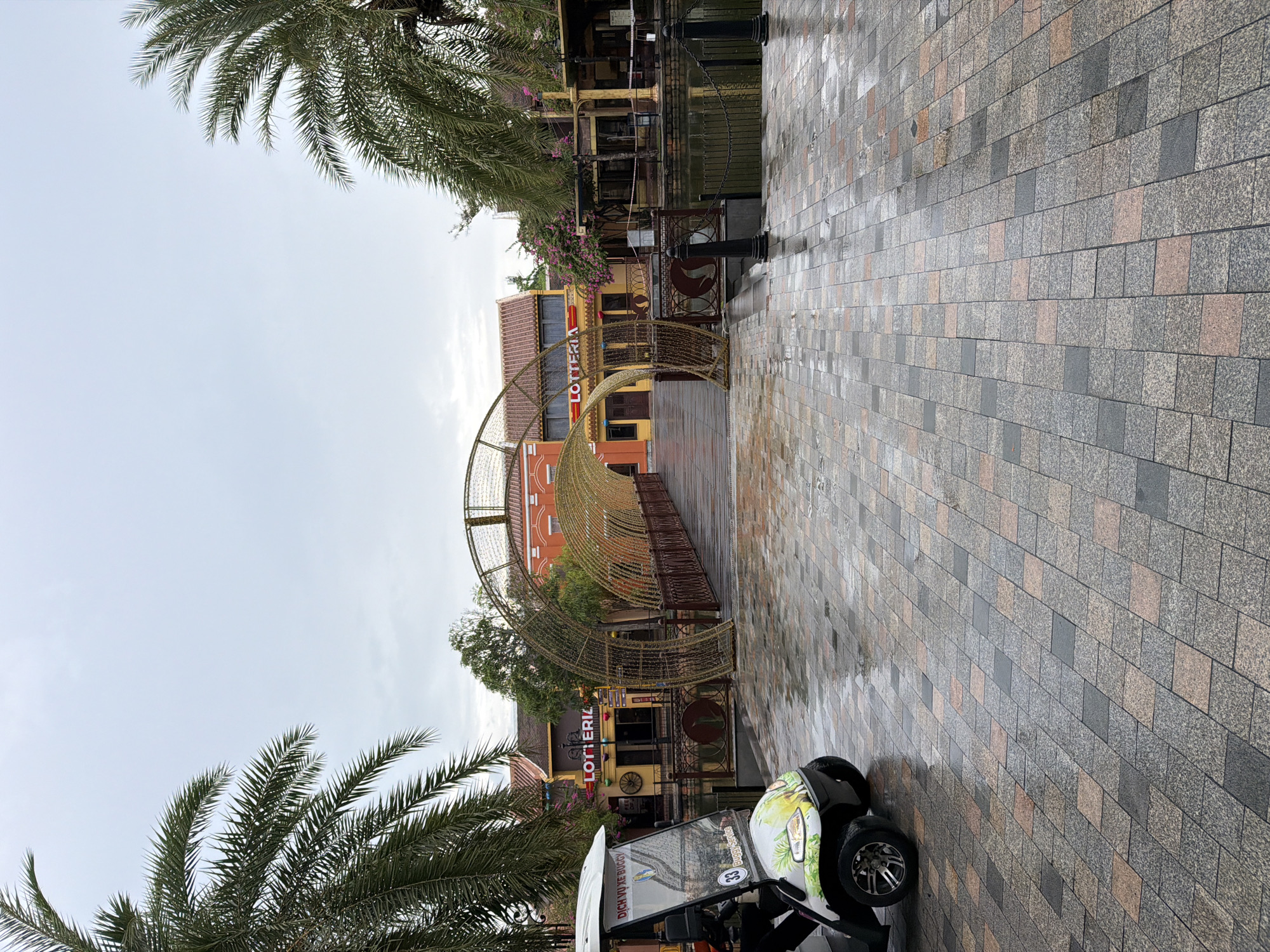 The image depicts a street scene in Da Nang, Vietnam, likely in a tourist or entertainment area. In the foreground, a white golf cart is parked on a wet, patterned brick pathway. The cart has a colorful decal of a bird and green foliage on its side and displays the number "33" in a blue circle, along with text that includes "DỊCH VỤ BUGGY" and "Buggy Service."
Beyond the cart, a wooden bridge spans a body of water. The bridge is covered by an illuminated arched structure made of golden, mesh-like material, creating a tunnel effect. On either side of the bridge, buildings with orange and yellow facades are visible. One building prominently features a red "LOTTERIA" sign. Palm trees line the sides of the scene, with their fronds reaching towards a overcast sky. The brick pathway is wet and reflects the ambient light, indicating recent rain. The overall impression is a festive and possibly commercial area, designed to attract visitors.