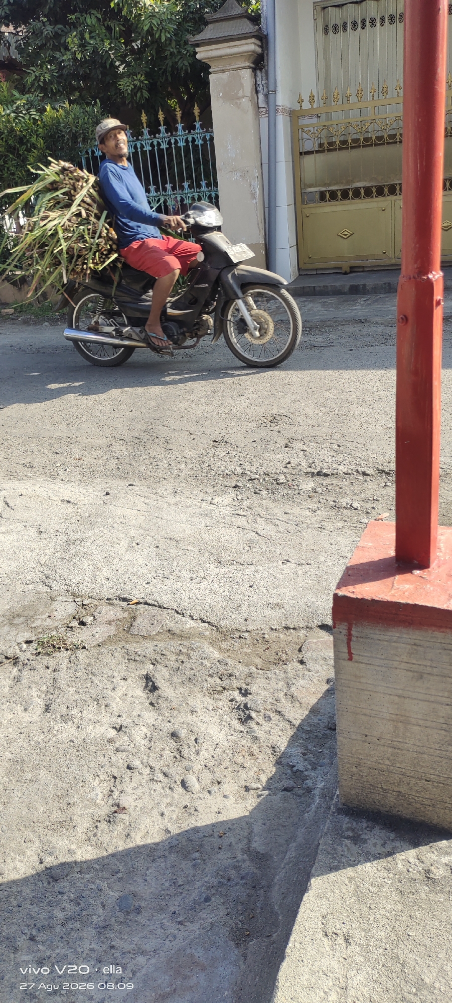An Indonesian man wearing a blue long-sleeved shirt and red shorts is riding a motorcycle laden with a large bundle of sugarcane stalks. He is wearing a camouflage-patterned cap and flip-flops. The motorcycle is dark-colored and appears to be a standard street bike. The man is looking towards the right side of the frame. The scene takes place on an asphalt road with some cracks and debris. Behind him, a beige building with a golden gate and ornate fencing is visible. Lush green trees line the background. A red cylindrical pole with a concrete base stands in the foreground to the right, casting a shadow across the road. The image was taken outdoors during the daytime.