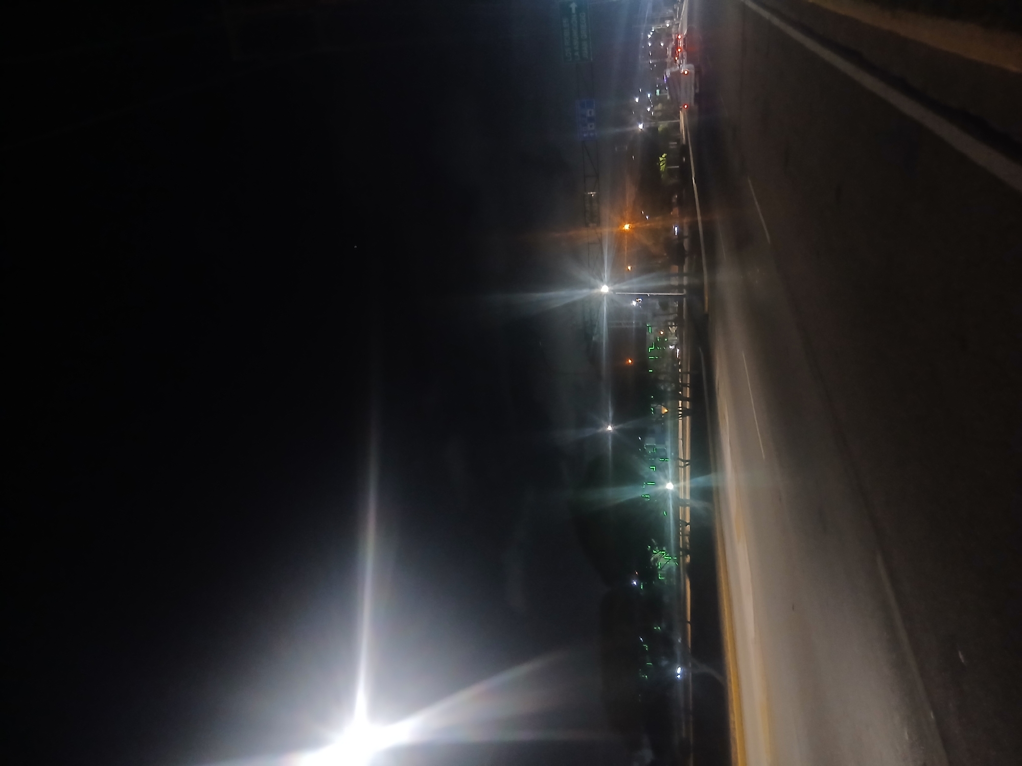 The image shows a night scene in Pampatar, Venezuela. On the right side is a road with lane markings and what appears to be a guardrail on the left side of the road. On the other side of the guardrail, there are streetlights and other illuminated structures, possibly buildings or urban features. The left part of the image is largely dark, with a bright light source near the bottom edge and various city lights visible in the distance. There are blurred vehicles in the upper portion of the frame.