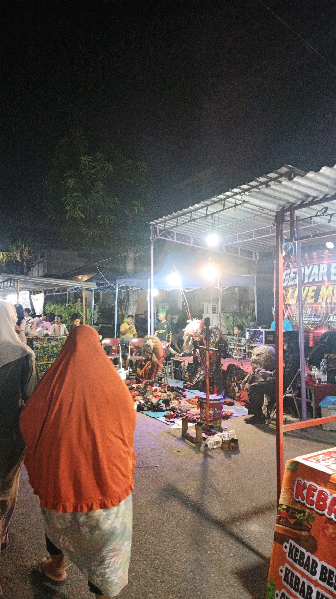 The image depicts an outdoor night scene in the city of Surakarta, Indonesia, likely at a street performance or festival. In the foreground, a person is seen from behind, wearing an orange headscarf and a patterned skirt. To the left, another person is partially visible, wearing a grey headscarf.
The background features a stage area under a canopy, illuminated by bright lights. On the stage, several individuals appear to be performers, some wearing elaborate costumes that include masks or headdresses. There are also musical instruments and sound equipment present. A crowd of spectators is gathered around the stage, some seated and others standing. In the far right of the image, a food stall is visible, with an orange banner displaying the word "KABAB." The ground is paved and appears to be a street or open area. The overall atmosphere suggests a lively cultural event.