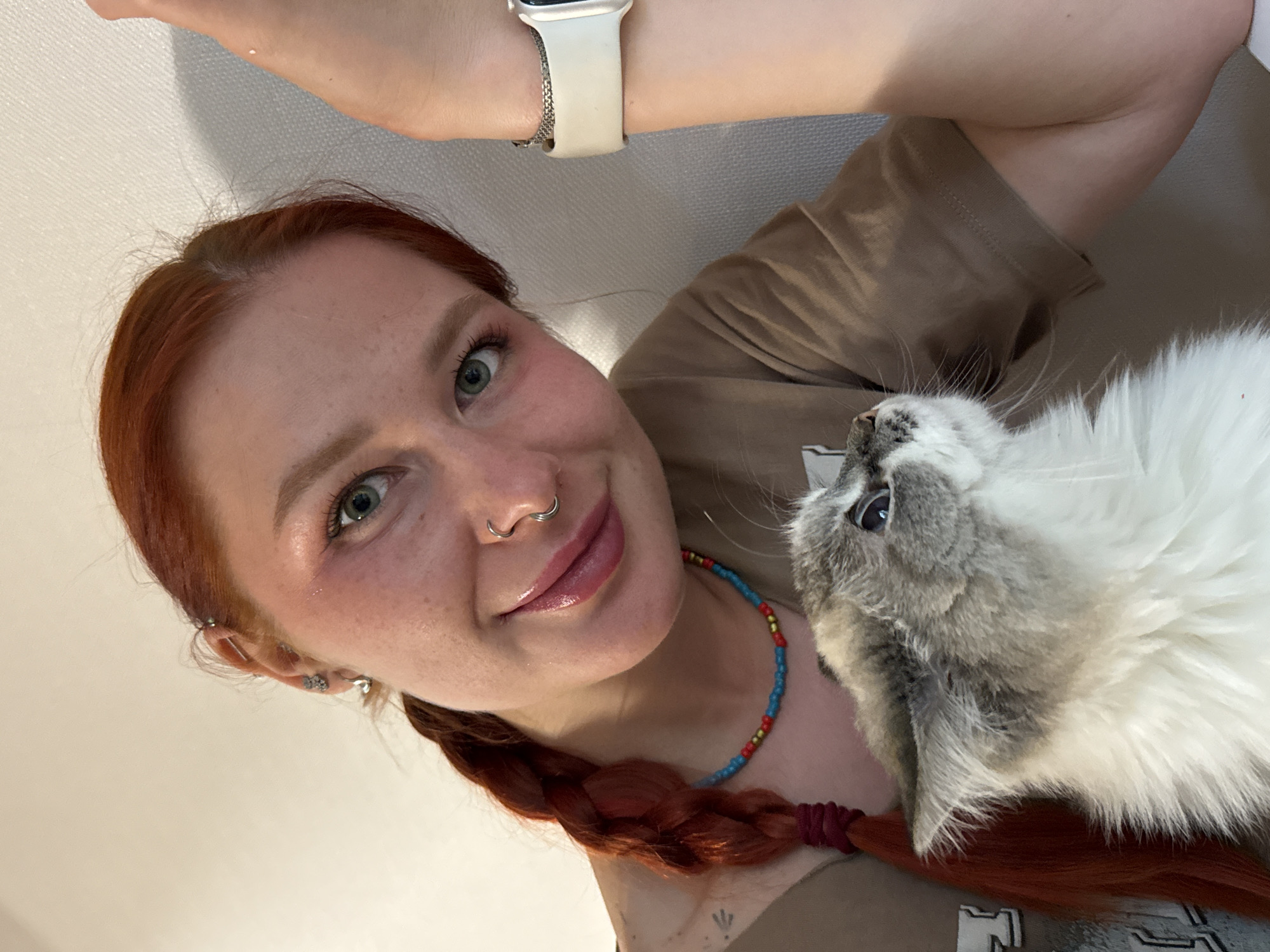 A selfie shot shows a light-skinned woman with auburn hair styled in a braid. She is looking into the camera, smiling, with a beaded necklace around her neck and multiple piercings on her left ear and nose. She's wearing a beige t-shirt and a white smartwatch. Positioned closely to her is a fluffy grey and white cat, its head near her shoulder. The background is a plain white wall. There is no evidence of the city of Rostov-on-Don, Russia.