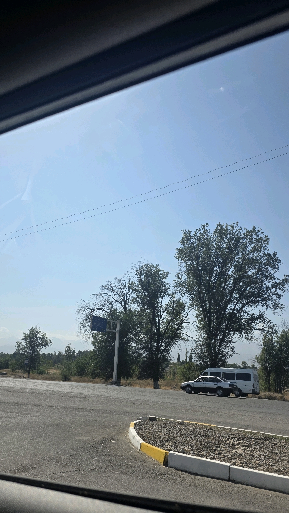 The image depicts a road intersection in the city of Tokmok, Kyrgyzstan, on a clear, sunny day. A light-colored sedan is stopped at the intersection, positioned behind a white van. The road surface is asphalt, with a curb marked by white and yellow paint. Scattered gravel and small rocks are visible within the curb. A white signpost with blue lettering stands near the road, indicating directions to various locations. Lush green trees are abundant in the background, with some appearing to be deciduous. In the distance, faint mountains can be seen under the bright blue sky. The image appears to be taken from inside a vehicle, as evidenced by the dark frame at the bottom and top of the photograph.