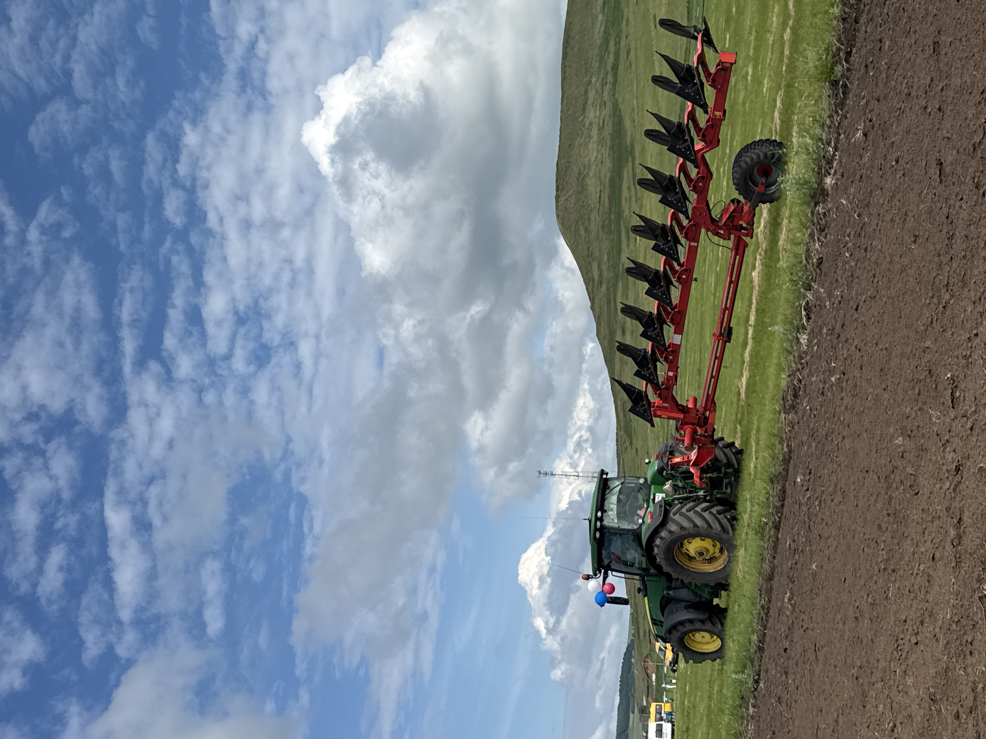 A green tractor with a red multi-furrow plow is in a field under a cloudy blue sky. The tractor is on the left side of the image, facing right, with its plow attached and extending to the right. The field is green grass, with a patch of tilled brown soil in the foreground. In the background, there is a rolling green hill and a few distant trees. A white van and a yellow vehicle are visible on the far left of the image. The scene appears to be in a rural area, possibly a farm or agricultural exhibition. The location is not identifiable as Nurkeevo, Russia based on the provided visual information.