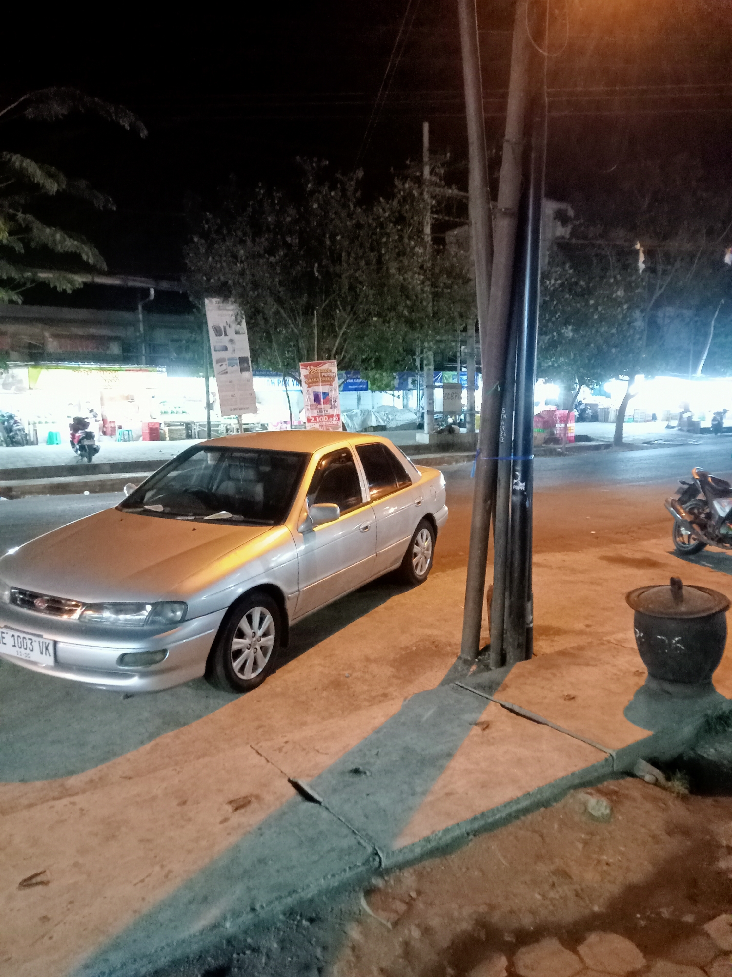 The image depicts a nighttime street scene in Mangunsari, Indonesia. A silver sedan is parked on the right side of the road, its headlights on. The car's license plate reads "BE 1003 VK". In the background, several shops with bright lights are visible, and a few motorcycles are parked in front of them. A large utility pole stands on the right side of the image, with wires attached. A dark-colored trash receptacle is situated at the base of the pole. The ground is paved with asphalt and concrete, and shadows are cast by the streetlights and the utility pole. Trees are scattered throughout the scene, their leaves illuminated by the artificial lighting.