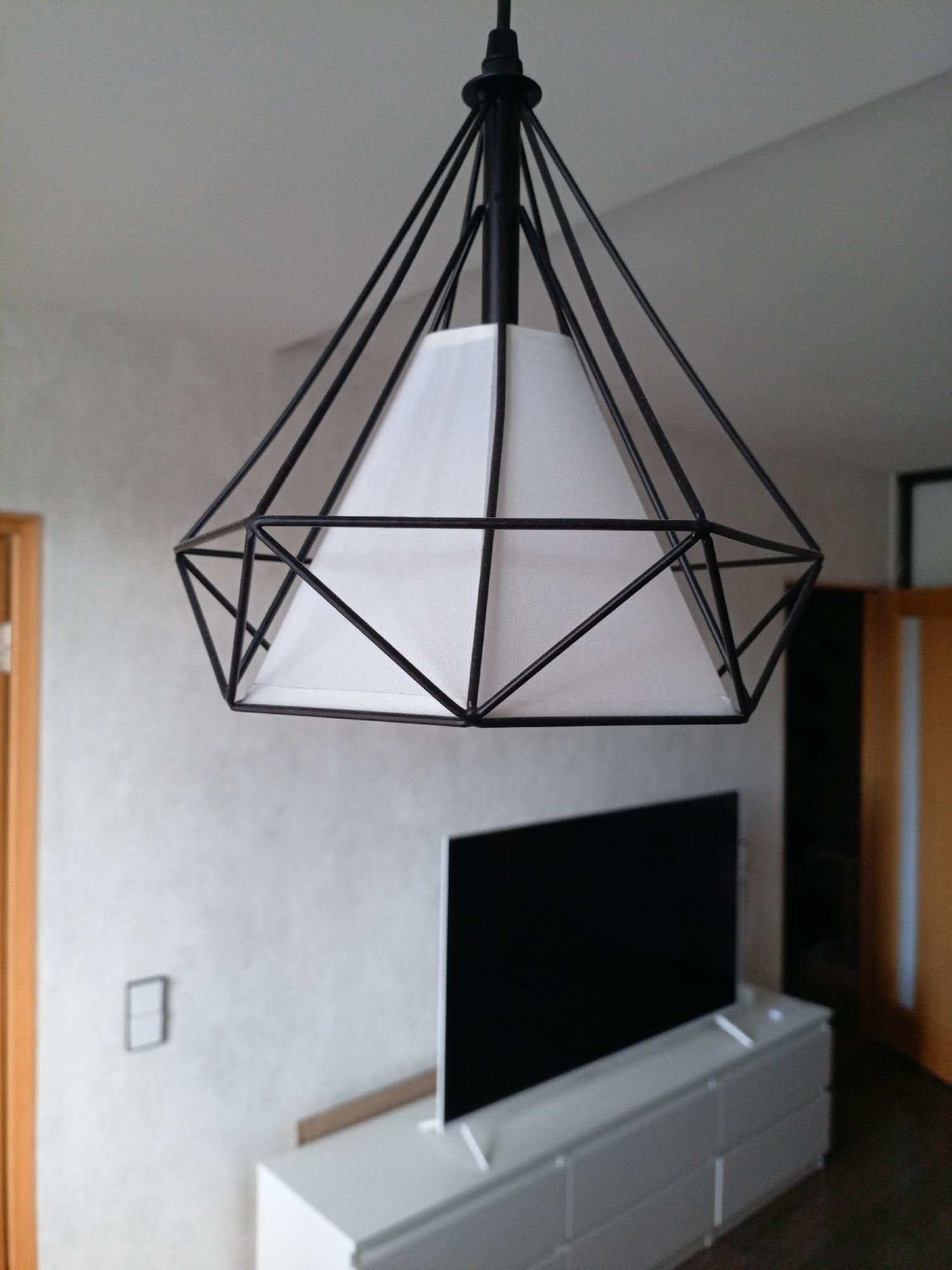 The image shows a modern living room interior. A black geometric pendant light fixture with a white fabric shade hangs from the ceiling in the foreground. Below the light, a black flat-screen television sits atop a white media console with drawers. The wall behind the television is textured and light-colored, with a small white electrical outlet cover visible. To the left of the television, a wooden door frame is partially visible. To the right of the television, a tall, dark cabinet and another wooden door with vertical white strips are present. The scene is well-lit, suggesting an indoor residential setting.