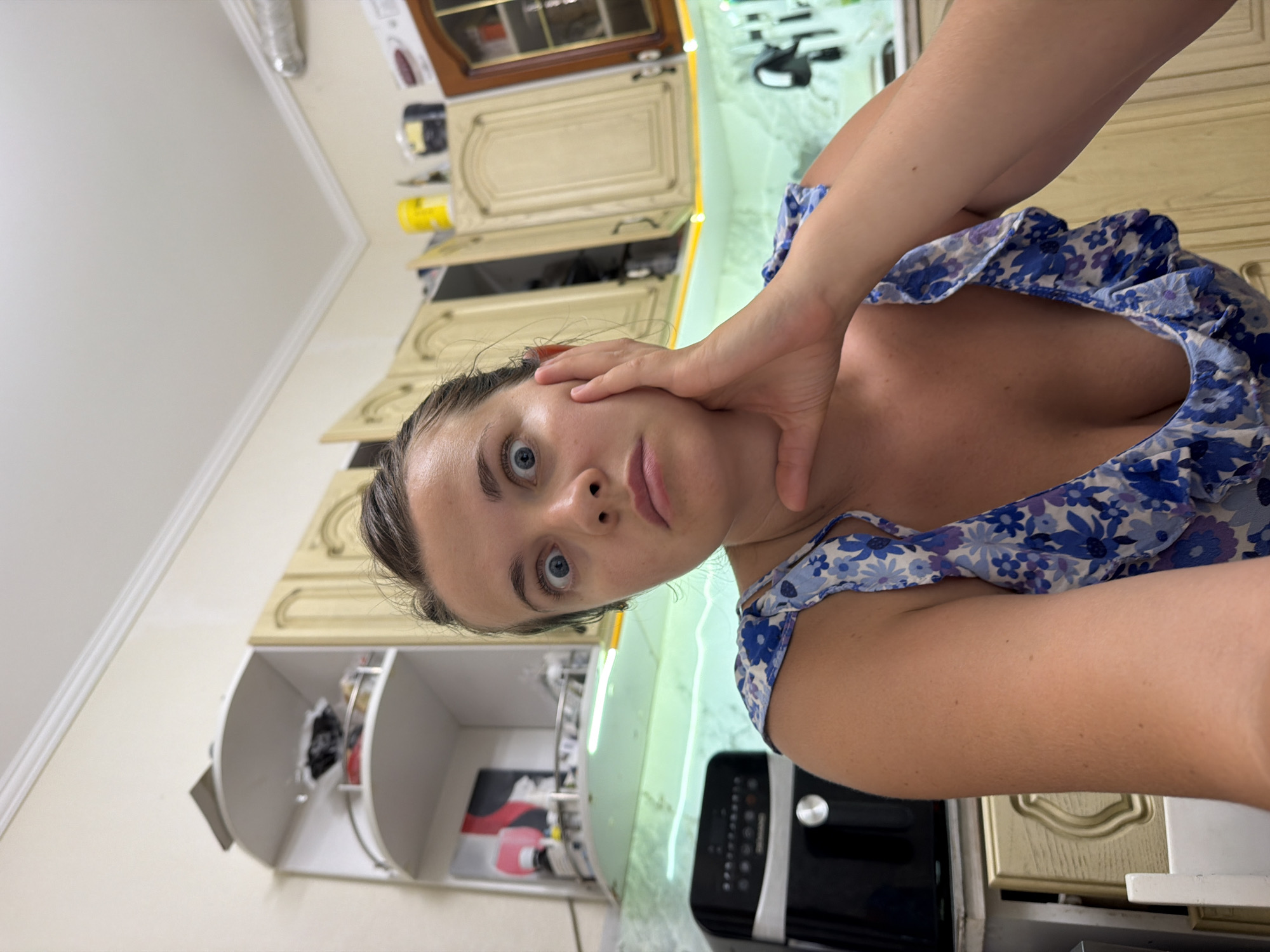 The image shows a close-up, waist-up shot of a woman with wide eyes and a surprised expression, her hand on her cheek. She is wearing a blue floral dress with ruffles. The background appears to be a kitchen, with light-colored cabinets, shelves with items, and an air fryer visible. The overall lighting suggests an indoor setting, possibly a home. The image does not contain any visual information to suggest the location is Sochi, Russia.