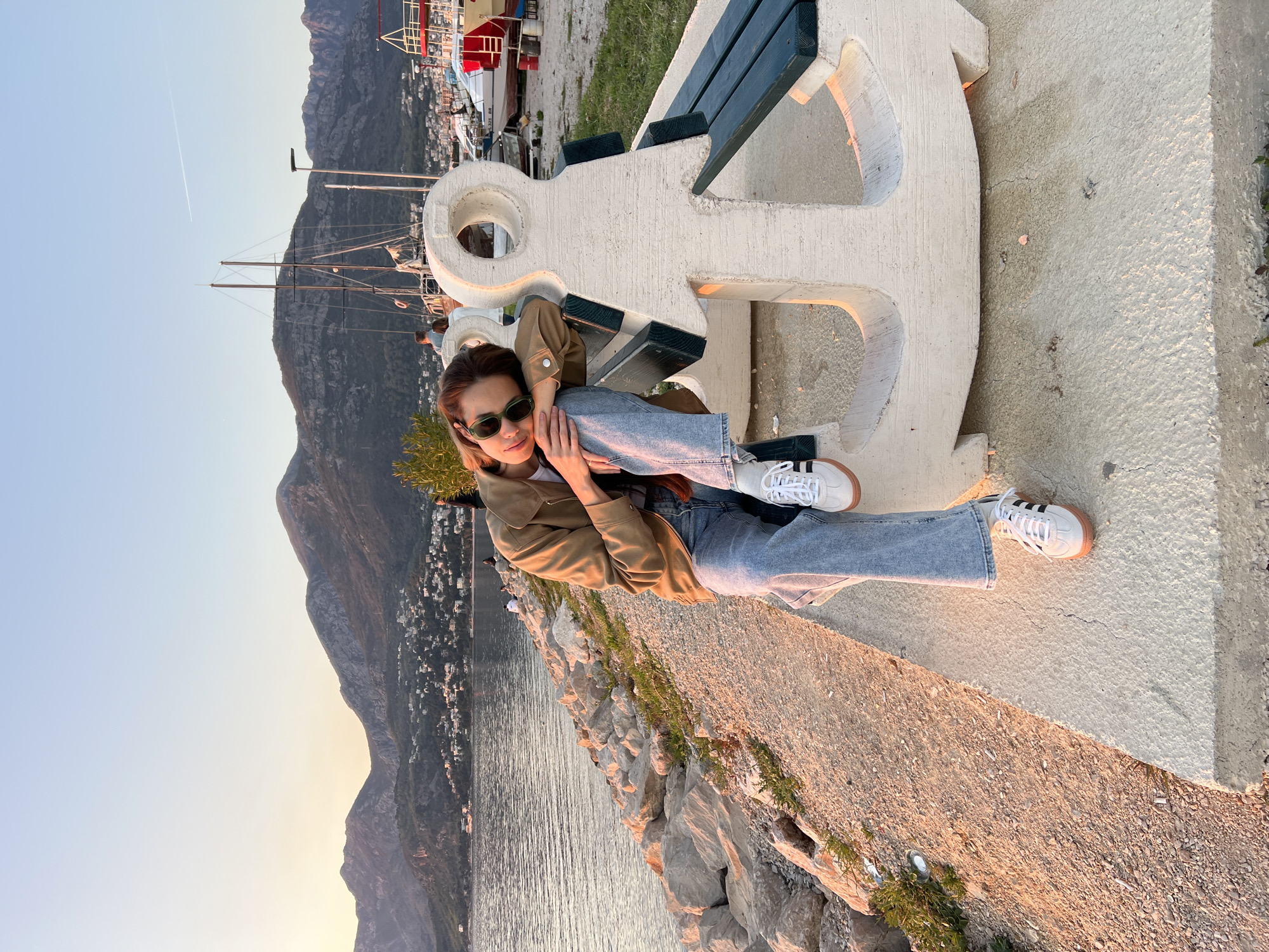 A woman is lying on a concrete anchor-shaped bench, positioned along a rocky shore near the Adriatic Sea. The mountains rise in the background, indicating a coastal location. A line of buildings are visible along the shore. The setting appears to be at dusk or dawn, with soft, warm light casting shadows. The woman is wearing a khaki jacket, jeans, and white sneakers with black stripes. She is wearing sunglasses and looking towards the viewer. The bench is white and includes a section of dark green slats. Some boats and masts are visible in the background. This location is Bar, Montenegro.