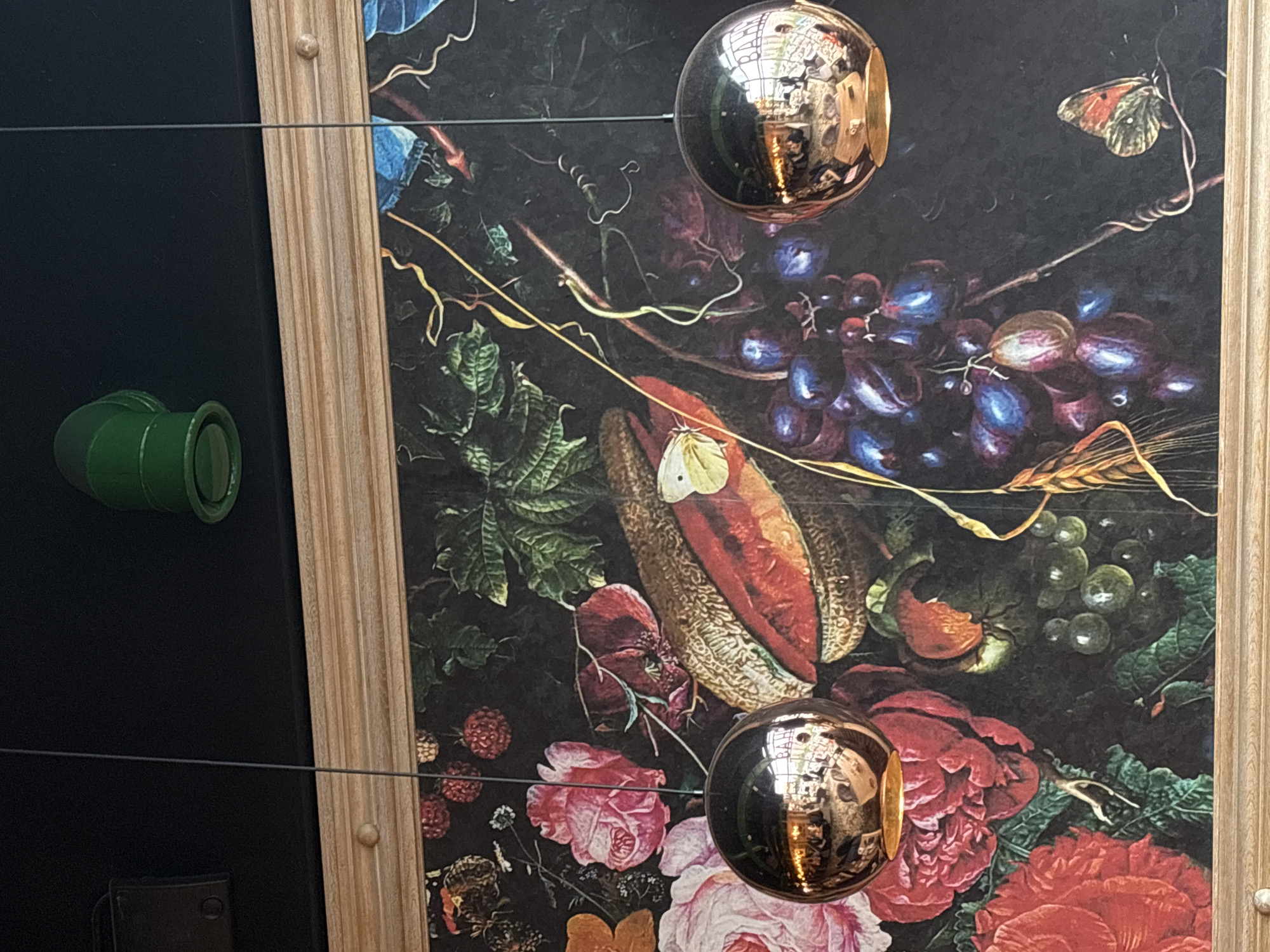 A framed still-life painting of flowers, fruits, and insects is visible. Two reflective spherical ornaments hang in front of the painting, reflecting light and the surrounding environment. A green, cylindrical object is visible on the left side of the frame, next to a dark surface. The presence of a wooden frame and reflective orbs suggests an interior, possibly a gallery or exhibit space in Saint Petersburg, Russia.