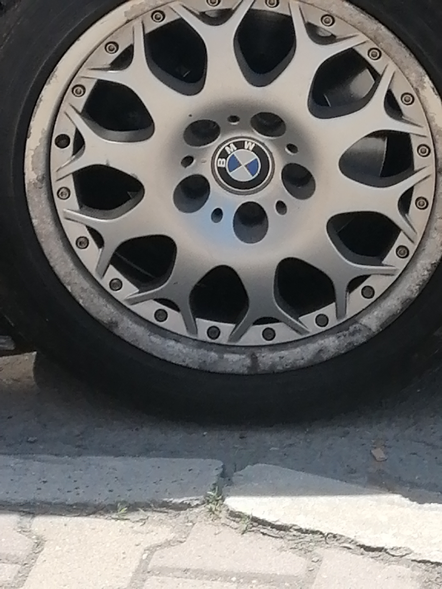 The image shows a BMW car wheel close-up. The wheel is silver with a BMW logo in the center. The tire is black. The wheel shows signs of wear and tear. The background includes a sidewalk with concrete blocks. The location cannot be determined based on the image, but the user specified that it is Sofia, Bulgaria.