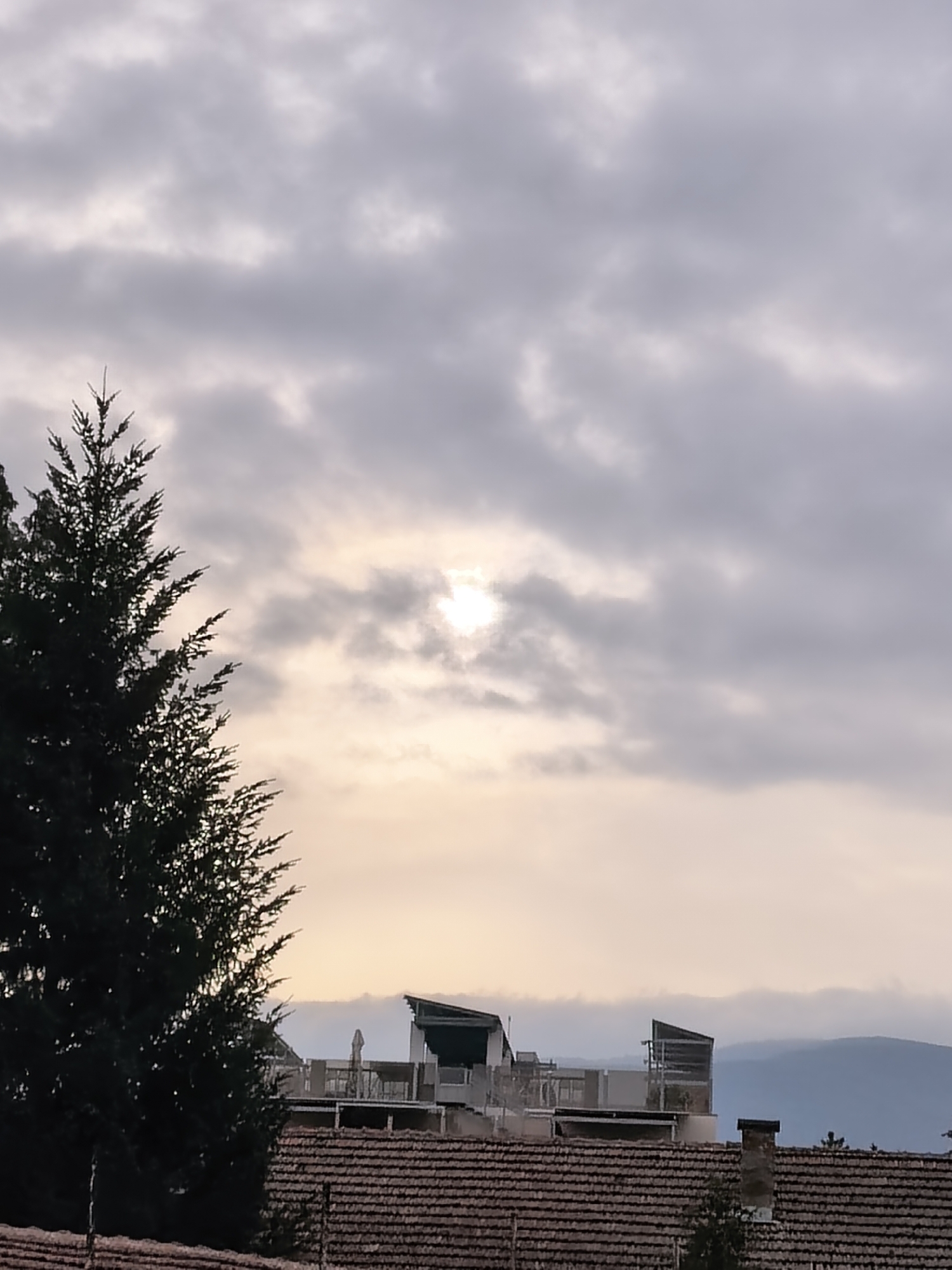 The image captures a cloudy sky in Unknown, Unknown, with the sun partially obscured by clouds. A tall evergreen tree is visible on the left side of the frame. In the lower portion of the image, there is a tiled roof of a building with a structure under construction on top. In the background, distant hills are seen.