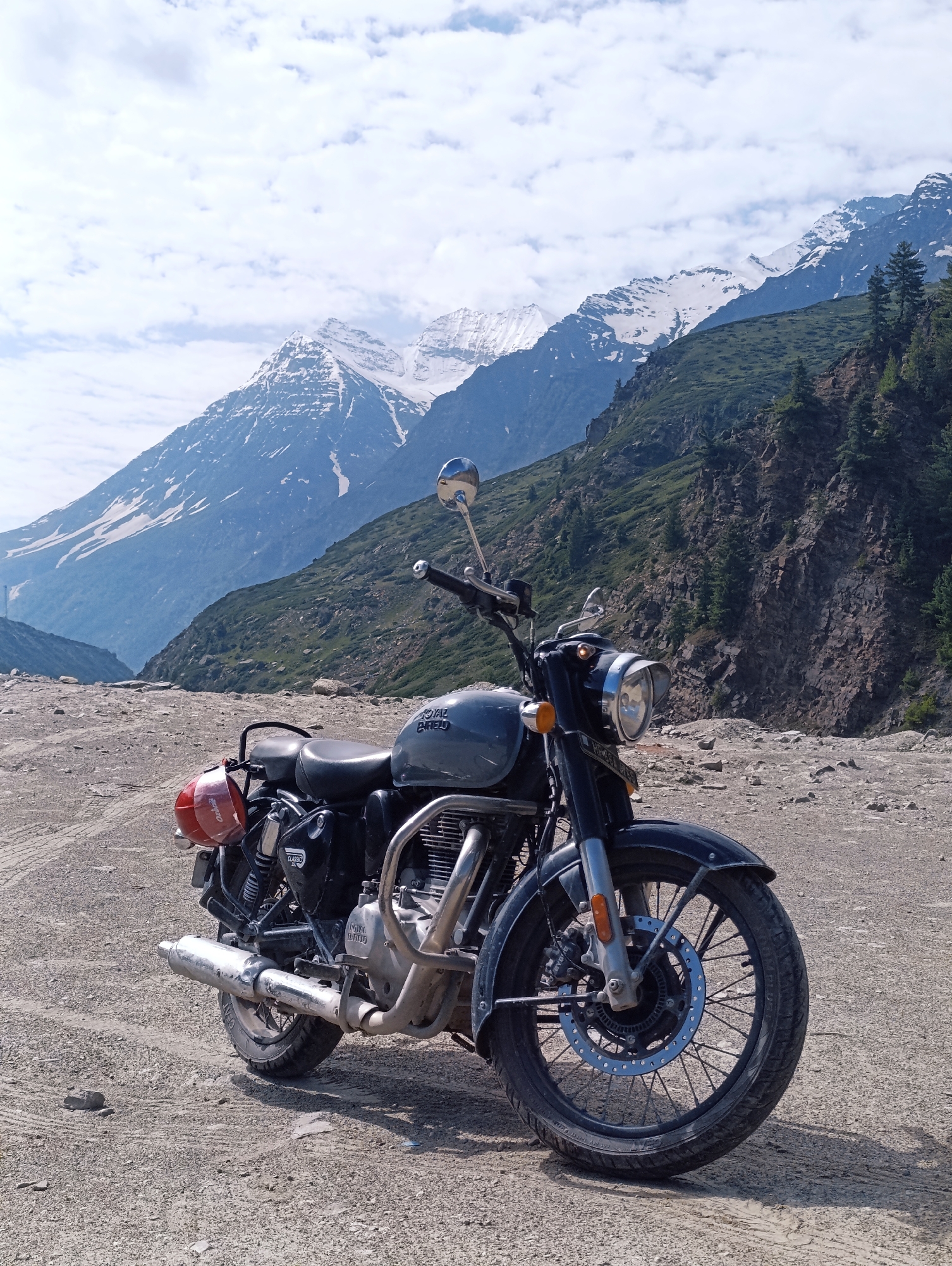 A dark gray Royal Enfield motorcycle is parked on a gravelly, dusty road in the mountainous region of Sissu, India. The motorcycle is positioned facing slightly to the right, with its front wheel closer to the viewer. The bike features a red rear fender light, a prominent exhaust pipe, and a silver engine guard. In the background, a vast mountain range dominates the scene. The lower slopes of the mountains are covered in green vegetation and rocky outcrops, while the higher peaks are capped with snow. The sky above is a mix of light blue and white clouds. The overall environment suggests a remote, natural setting in the Himalayas.
