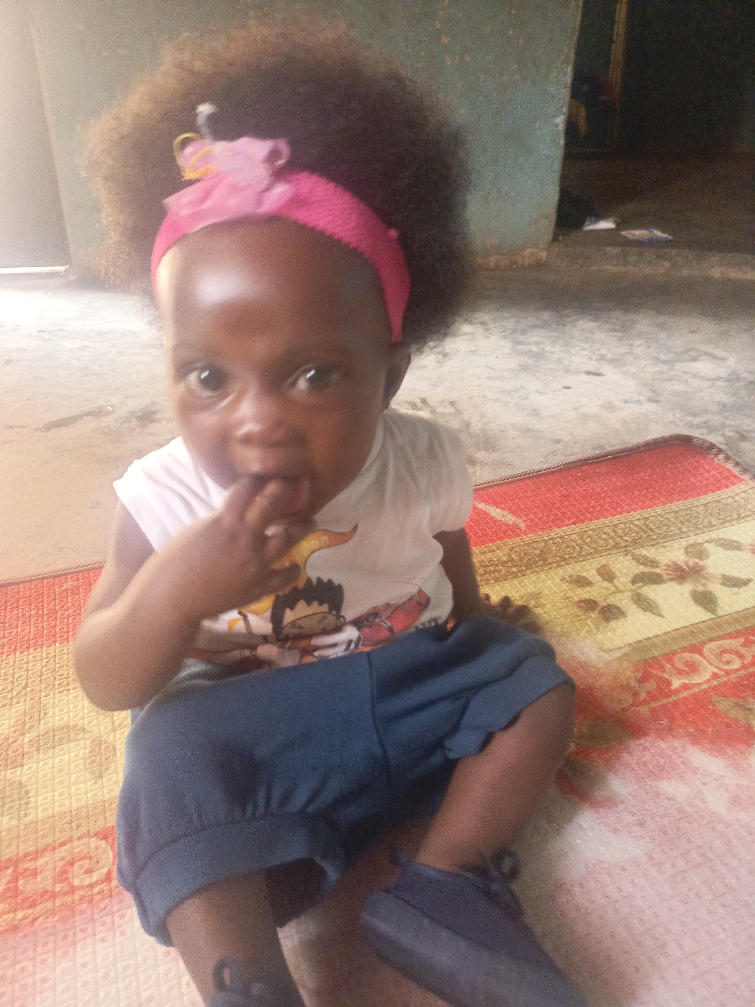 A young child is seated on a patterned mat. The child wears a white shirt with a cartoon image, blue shorts, and matching shoes. A pink headband adorns the child's hair. The child has fingers in their mouth. The background shows a concrete floor and wall, consistent with a domestic interior. The image was likely taken in a home within Akwanga, Nigeria, given the prompt.