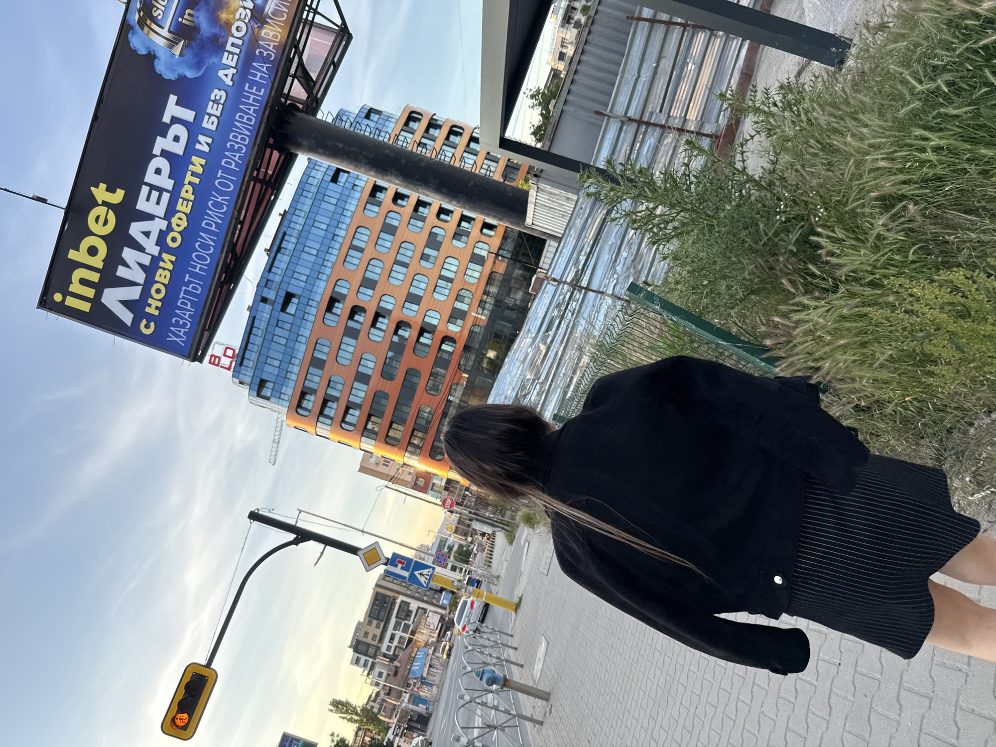 The image shows an outdoor scene in Sofia, Bulgaria, at what appears to be dusk or early evening. Dominating the left side of the frame is a large billboard advertising "inbet", the text reading "ЛИДЕРЪТ С НОВИ ОФЕРТИ И БЕЗ ДЕПОЗИ ХАЗАРТЪТ НОСИ РИСК ОТ РАЗВИВАНЕ НА ЗАВИСИ". A building with orange and blue-tinted glass windows is visible in the background, angled slightly to the right.
To the right of the billboard, the upper-right portion of the frame includes overgrown green vegetation. A corrugated metal structure is located just behind the vegetation.
In the foreground, a person wearing a black jacket and a striped skirt is walking along a paved sidewalk. Only their back is visible.
Further into the background, there is a street scene with street signs, traffic lights showing a yellow signal, other buildings, and parked cars. The street and surrounding area suggest an urban environment. The presence of bicycle racks near the sidewalk indicates a pedestrian and cyclist-friendly space.