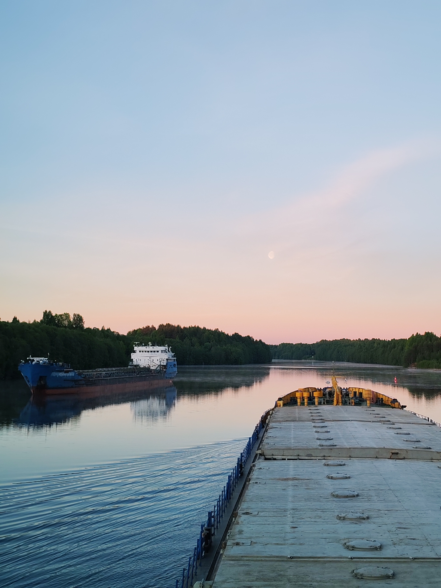 The image captures a serene river scene during twilight, likely at either sunrise or sunset, in Volokov Most, Russia. The perspective is from the deck of a cargo vessel, with its front deck, featuring numerous hatch covers and yellow machinery, visible on the right side of the frame. Blue railings line the left edge of this vessel's deck.
In the midground, another large cargo ship, painted blue with a white superstructure, navigates the calm, reflective waters of the river, moving in the opposite direction. Its clear reflection is visible in the water, along with the gentle wake from the foreground vessel.
The river is bordered by dense green forests on both banks, suggesting a natural or semi-natural waterway. A subtle red buoy can be seen in the distance on the right bank. The sky transitions from a soft pale blue at the top to a warm pink and orange near the horizon, illuminating a distinct, bright white moon positioned in the upper right quadrant. No people or discernible text are visible in the tranquil scene, which focuses on the peaceful passage of vessels through a natural landscape.