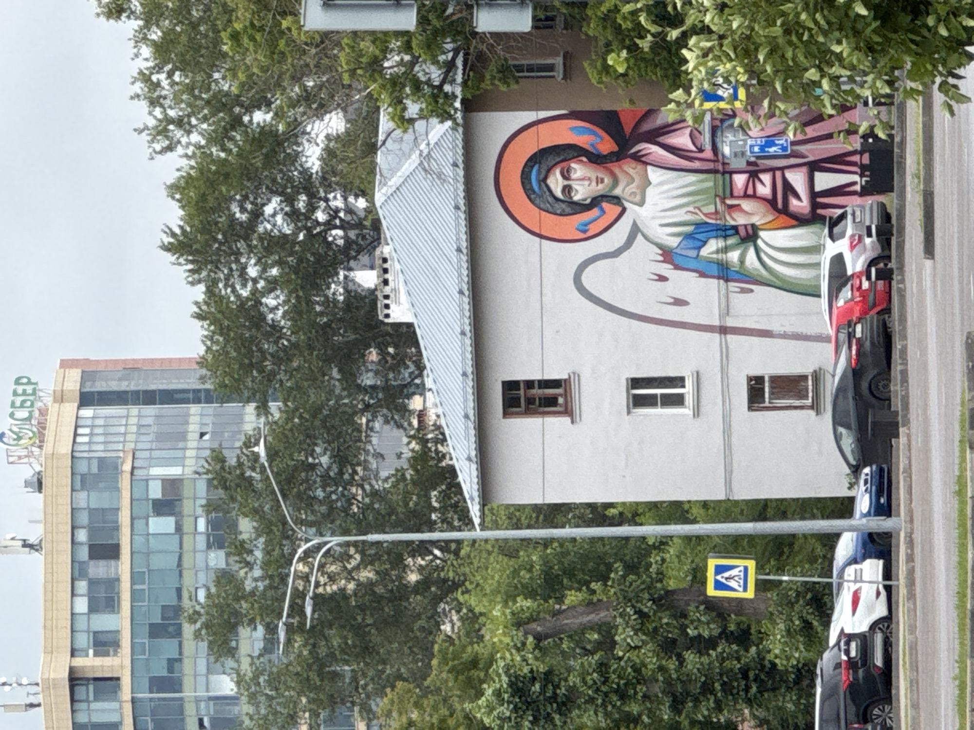 The image shows a mural of an angel painted on the side of a light-colored apartment building. The building has a corrugated metal roof and several windows. In front of the building, there is a street with several parked cars and a pedestrian crossing sign. Behind the apartment building, there is a modern glass-fronted office building with the logo "СБЕР" (Sberbank) visible at the top. Trees surround the buildings. The scene is likely in Nizhny Novgorod, Russia, given the presence of the Sberbank logo and the architectural style.