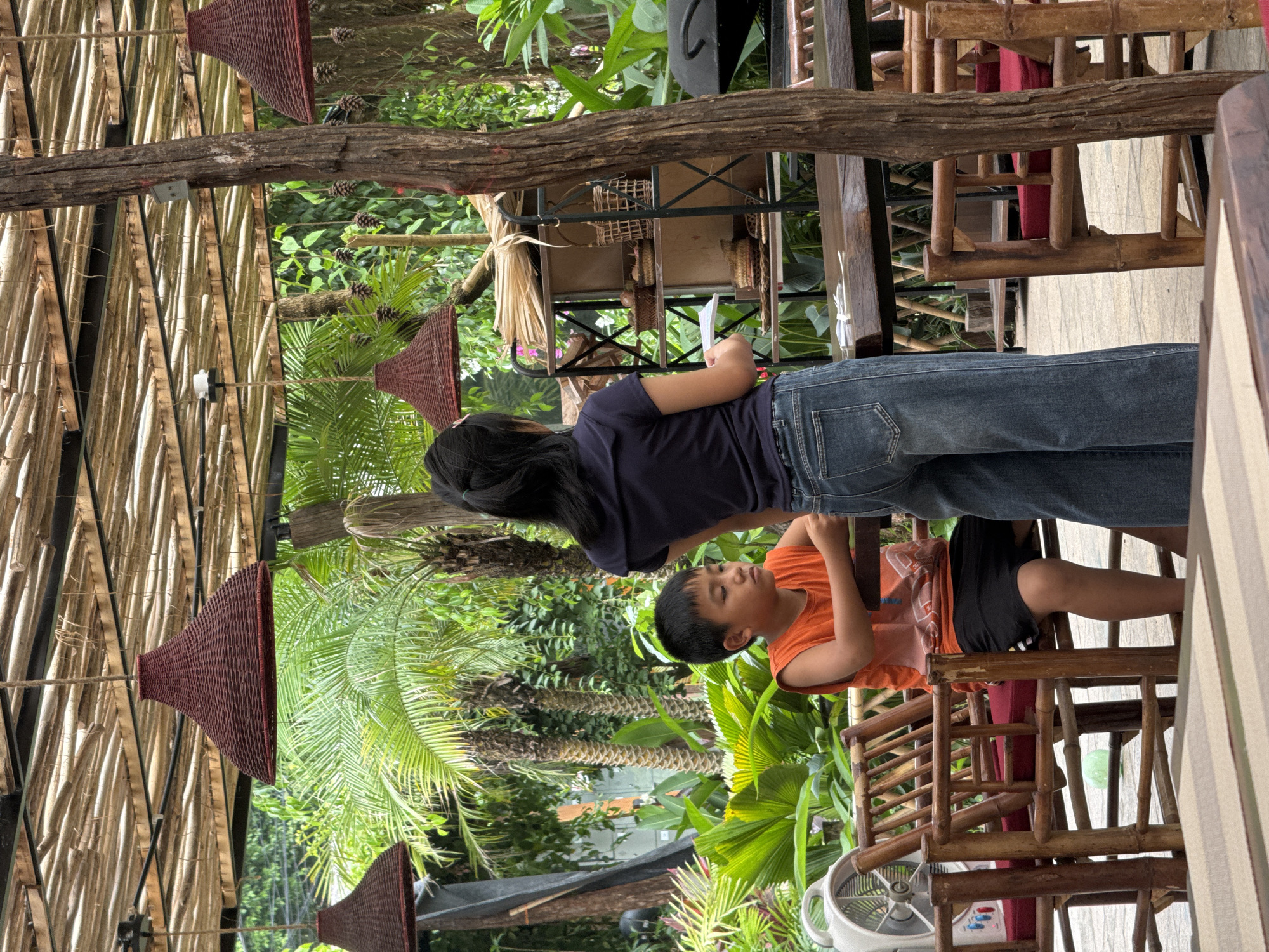 This is a medium-shot, eye-level photograph taken outdoors in a tropical setting, possibly a restaurant or cafe, in Nam Hoa Lư Ward, Vietnam. The scene is partially covered by a rustic bamboo roof supported by wooden beams and posts. Hanging from the roof are three woven, conical pendant lamps with a dark red hue.
In the foreground, two children are present. On the left, a young boy, appearing to be around 7-10 years old, sits facing the left of the frame. He is wearing an orange sleeveless shirt and black shorts, and his arms rest on a wooden table. His expression appears somewhat pensive. To his right, and slightly behind him, stands a girl, likely a teenager, with her back to the camera. She is wearing a dark blue t-shirt and loose-fitting denim jeans. She holds a white object, possibly a menu or paper, in her right hand and appears to be interacting with the boy or looking towards something out of frame.
The environment is lush with tropical vegetation, including palm trees and large, vibrant green leaves, suggesting a warm climate. To the left of the boy, a white electric fan with a circular base is visible. The furniture consists of rustic, light-colored bamboo chairs with red cushions, and wooden tables. In the background, there is a multi-tiered shelf unit made of dark metal and wood, displaying various woven baskets and items. Further in the background, a dark object, possibly a grill or part of another table, is partially visible. The ground appears to be paved with light-colored tiles. The overall impression is a relaxed, outdoor dining or gathering space amidst abundant greenery.