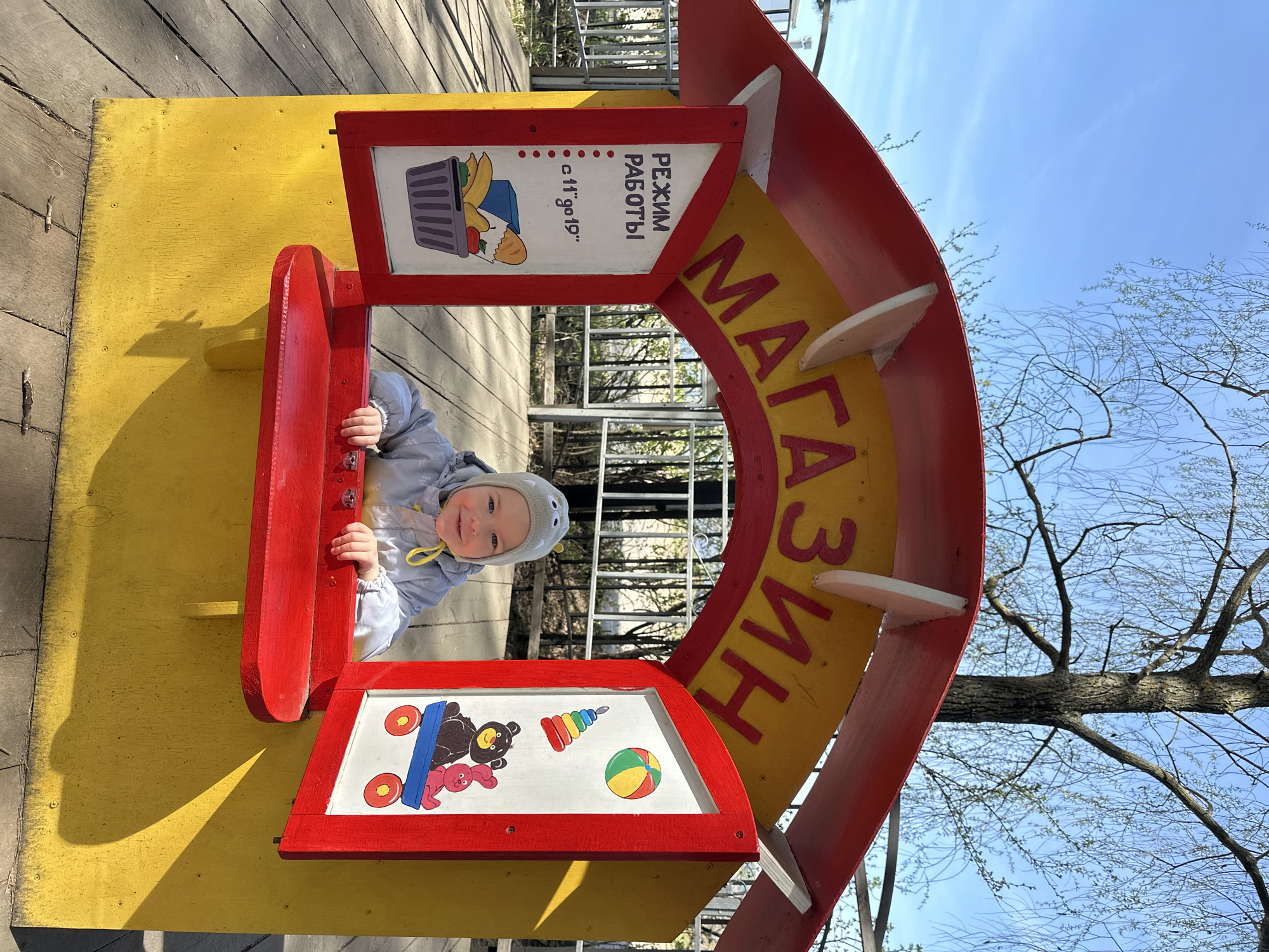 The photograph captures a young child, possibly a toddler, playing inside a brightly colored structure that resembles a store or shop facade at a playground. The child is fair-skinned with light hair, wearing a gray hooded jacket and matching hat with small decorative protrusions resembling eyes. The child is smiling and looking directly at the camera.
The structure is painted in bold primary colors, with red and yellow being dominant. It has a window or opening where the child is positioned, and a sign that reads "МАГАЗИН" (MAGAZIN), which translates to "Store" in Russian. Additional signage is present, featuring the phrase "РЕЖИМ РАБОТЫ" (REZHYM RABOTY), meaning "Working Hours," and a depiction of a grocery basket. Another sign shows pictures of toys.
The background consists of a metal fence or railing, trees with sparse foliage indicating either early spring or late autumn, and a clear blue sky. The scene appears to be a public park or playground. Based on the information in the prompt, the location is presumed to be Chaikovsky, Russia.