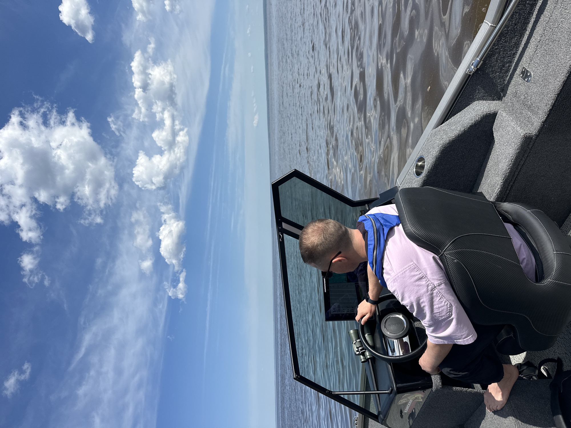 A person wearing a life vest and a light purple shirt operates a boat on water. The boat's instrument panel is visible, and the sky is visible in the background. The location cannot be confirmed as Novyi, Russia.