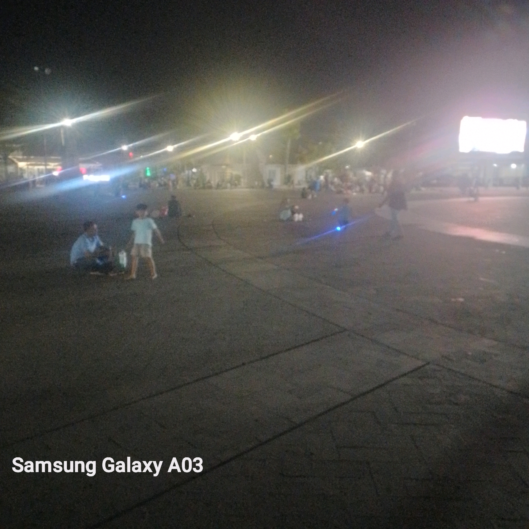 In Indramayu, Indonesia, a nighttime scene unfolds in a public square, illuminated by bright streetlights and a large, glowing billboard in the background. The ground is paved with dark asphalt, marked with light-colored lines indicating pedestrian pathways or parking spaces. In the foreground, a man in a blue shirt and dark pants sits on the ground, facing a young boy standing nearby in a light blue t-shirt and beige shorts. The man appears to be attending to something on the ground, possibly a bag or a picnic spread. The boy is looking towards the man, with his arms slightly outstretched. In the mid-ground, other individuals and small groups are scattered across the square, some appearing to be seated or standing. The overall atmosphere suggests a casual gathering or a break in an evening outdoor activity, with the presence of the billboard indicating a commercial or entertainment area.