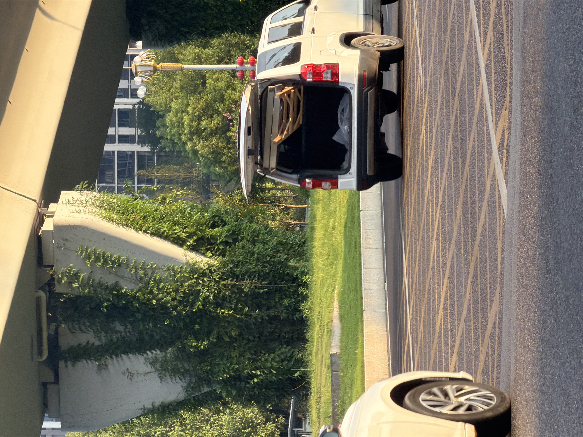 In Jiangjin, China, under a concrete overpass, a white SUV is parked with its rear hatch open, revealing a folded wooden item and plastic sheeting inside. The SUV is positioned on a road marked with yellow grid lines. To the left, the front quarter of a cream-colored car is visible. The environment features lush green vegetation climbing the overpass supports and surrounding the area. In the background, trees and a multi-story building can be seen. A traditional streetlamp with white globes and a decorative yellow base stands to the right of the SUV.