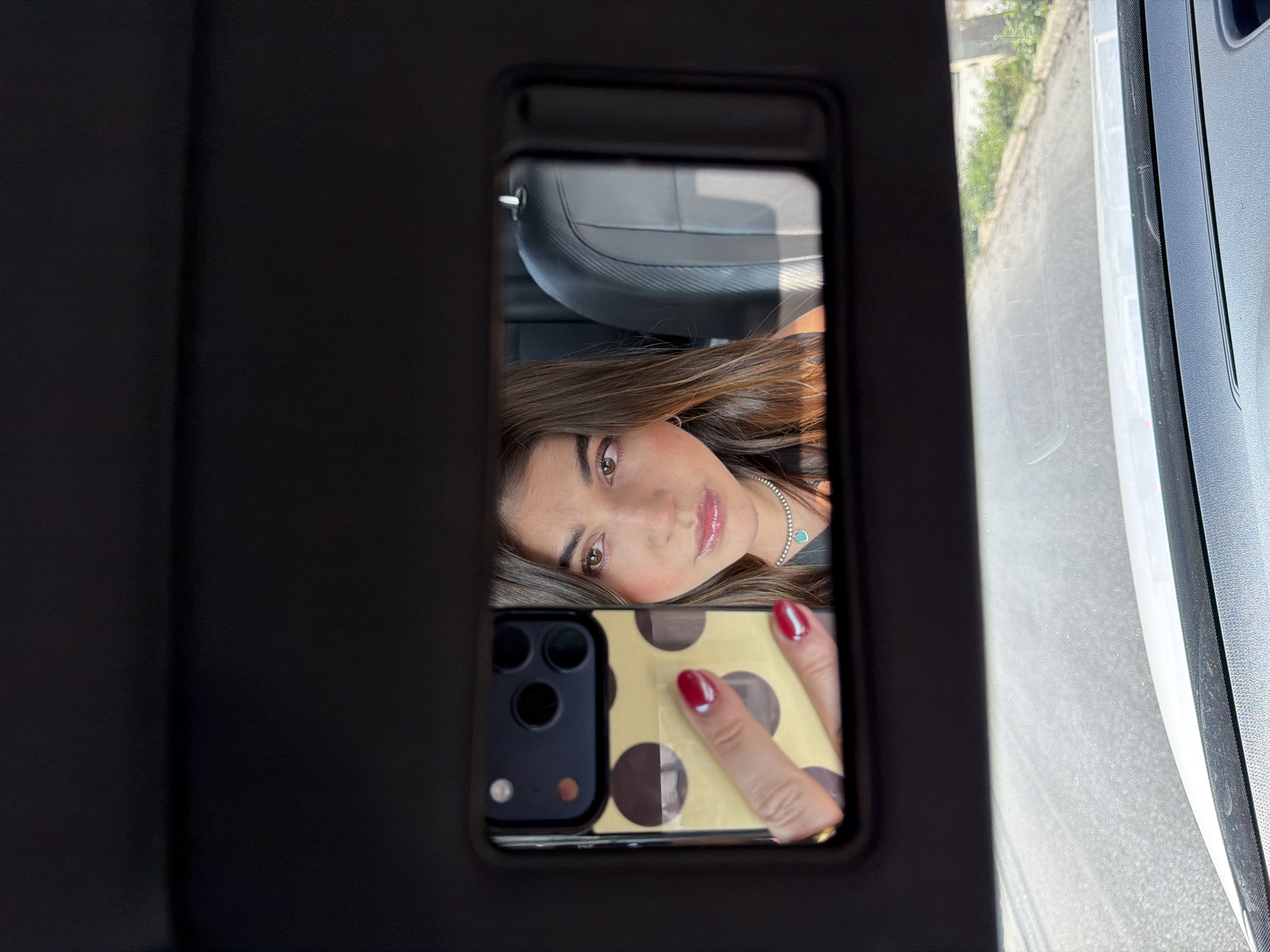 A close-up shot captures a young woman taking a selfie inside a vehicle, reflected in a car mirror. The image is oriented vertically, with the woman's head tilted. Her face, with brown eyes and brown hair flowing down, is central in the mirror's frame. She has a neutral expression, with glossy lips and light makeup. A silver-colored necklace with a small turquoise pendant adorns her neck.
Her right hand, adorned with red nail polish, holds a smartphone with a distinctive yellow or gold case featuring large brown polka dots. The multiple camera lenses of the phone are visible in the reflection.
The immediate surroundings within the mirror show the dark interior of the car, including a portion of a seat. To the right of the mirror, outside the car's window, a blurred view suggests a daytime street scene. Green foliage (bushes or trees) and what appears to be a paved road or sidewalk are visible, hinting at an urban or suburban environment. The lighting is bright, indicating it's daytime. No visible text is present in the image. The scene is set in San Bernardino Tlaxcalancingo, Mexico.