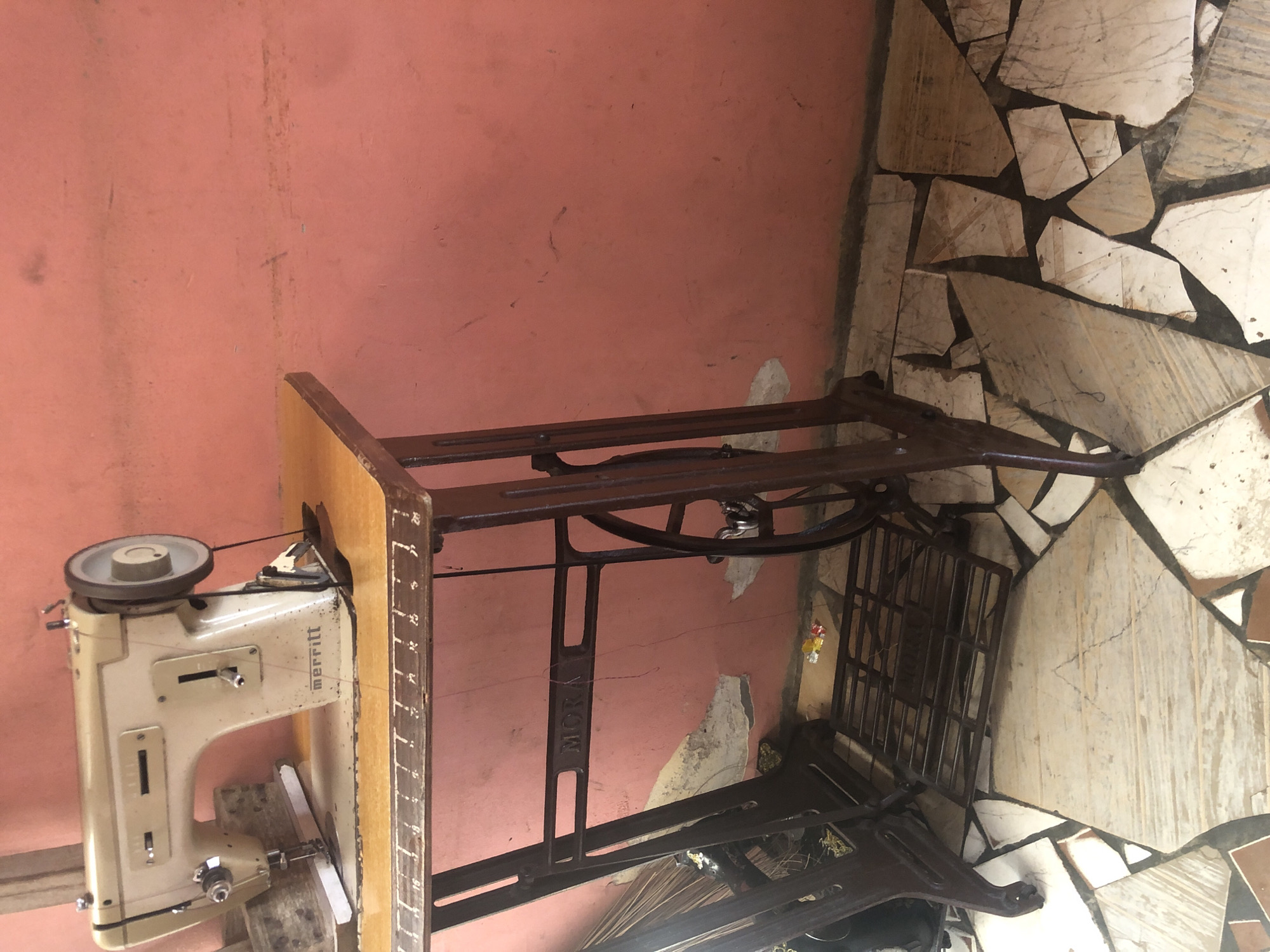 The image captures a vintage treadle sewing machine set up against a wall, likely indoors. The main subject is an off-white sewing machine, branded "MERRITT" on its side, mounted on a light-colored wooden table top. The machine has a large handwheel, various levers, and is threaded, indicating readiness for use. The wooden table features etched numerical markings along its front edge.
The sewing machine's table is supported by a dark, possibly cast-iron, metal stand. The name "MORAH" is visible vertically on one of the stand's upright supports. This stand incorporates a pedal mechanism at its base, characteristic of a treadle machine. The metal frame shows signs of age and wear.
The background features a worn, pinkish-peach colored wall with various dark marks and patches where paint or plaster has chipped away. The floor is covered with an irregular mosaic pattern, composed of broken or unevenly shaped light-colored tiles (beige, white, grey) with dark grout lines. Beneath the sewing machine stand, on the floor, some long, thin, dark objects (possibly sticks or broom bristles) are visible, along with a few small, colorful items hanging on a string or wire towards the right.
The scene depicts a functional workstation for sewing, but no individual is present or actively engaged in sewing. The visible text includes "MERRITT" on the sewing machine and "MORAH" on the metal stand. The overall impression is one of an older, well-used but functional setup in a practical indoor environment in Ado, Nigeria.