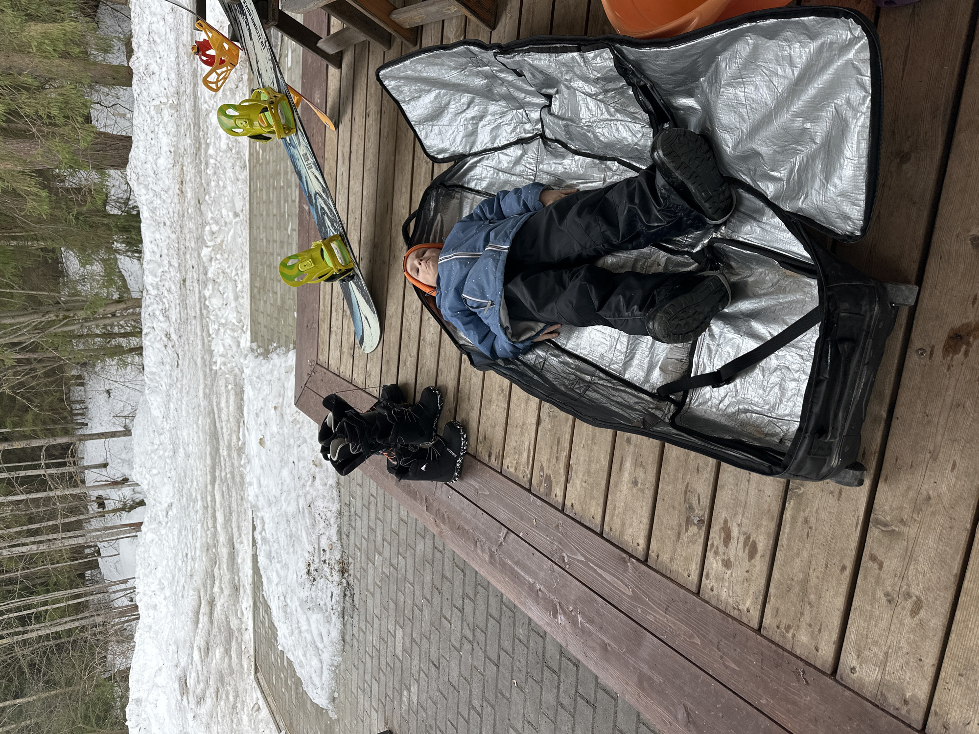A partially-filled ski travel bag lies on a wooden deck next to skis, boots, and snow in Прикамский, Russia. Inside the bag is a child lying on their back. They wear a blue jacket, black pants, black boots, and an orange knit cap. The ski bag is silver inside and black outside. A pair of skis with yellow bindings are nearby, leaning against a wooden structure. A pair of black ski boots are placed next to the ski bag. A pile of snow is visible next to a brick walkway and trees.