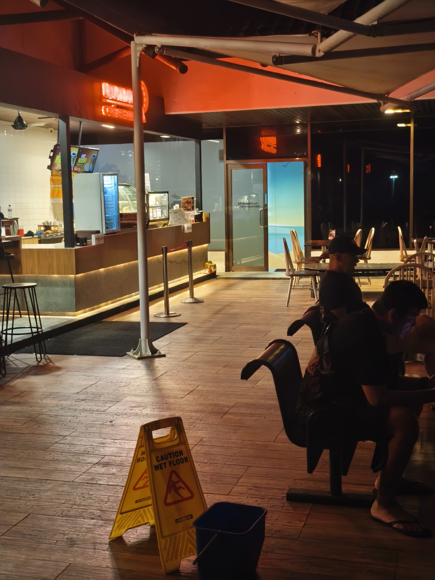 The image depicts the outdoor seating area of a cafe or restaurant at night. The scene is illuminated by warm, ambient lighting, including neon signs that spell out "RUNWAY CAFE". A wooden counter with illuminated trim is visible, along with display cases showcasing food items. Two people are seated at a table in the foreground, appearing relaxed as they engage with their phones. Another person, wearing a black t-shirt and cap, is seated in a unique chair, also seemingly absorbed in their device. A yellow "Caution Wet Floor" sign and a blue bucket are placed on the wooden floor, suggesting recent cleaning. The overall atmosphere is casual and inviting, with the warm lighting and comfortable seating creating a welcoming ambiance. The time of day is clearly night, and the lack of visible weather elements suggests clear conditions. The presence of outdoor seating and the general ambiance point to a relaxed dining or social setting, likely in a tropical or coastal location given the style of the establishment. The neon sign clearly indicates the establishment's name as "RUNWAY CAFE".