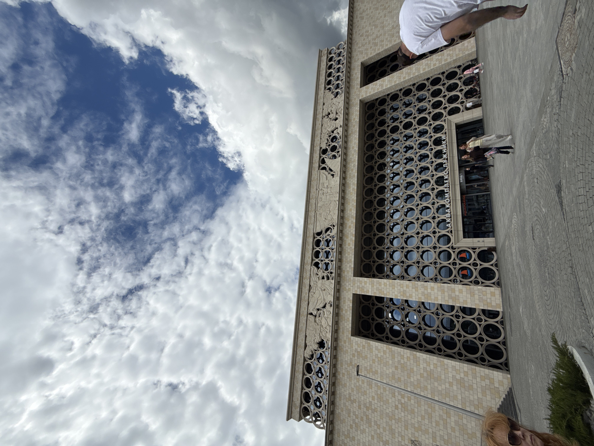 This image captures a low-angle view of a modern building with a distinctive facade, set against a dynamic sky filled with clouds. The building's exterior features a beige, tiled surface and a prominent section with a repeating pattern of circular openings, revealing dark interiors. To the left of this patterned section, a vertical band of intricate bas-relief carvings depicts figures and mythological scenes, adding an artistic element to the architecture.
Several people are visible on the paved area in front of the building. One individual, wearing a white t-shirt, is partially visible at the top right, with their legs extending downwards. Closer to the building entrance, a small group of people are gathered, some appearing to be posing for a photo or engaged in conversation. One person wears a light-colored dress, and others are dressed in darker clothing. A few more individuals are seen further down the sidewalk.
The scene appears to be taking place during the daytime, with a mix of bright light and shadow suggesting either early morning or late afternoon. The sky is partly cloudy, with dramatic white and grey clouds interspersed with patches of blue sky, indicating a somewhat overcast but likely mild weather condition.
Visible text on the building facade includes Cyrillic characters above the entrance, reading "АКВАКОМПЛЕКС ЛУЖНИКИ," which translates to "Luzhniki Aqua Complex." Another sign reads "ВХОД 1," meaning "ENTRANCE 1." The paving in front of the building is made of interlocking stones, with some circular patterns visible in the design. A small evergreen shrub is present on the right side of the frame. The overall impression is of a public space, possibly a recreational or cultural complex, with people enjoying the outdoors and the striking architecture.