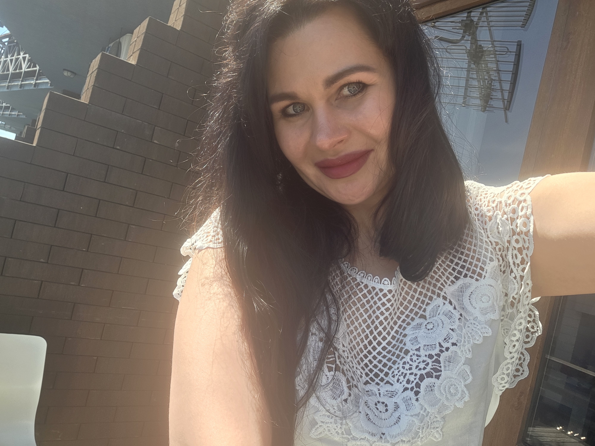A medium close-up selfie shows a woman with dark, wavy hair looking directly into the camera. She has light-colored eyes, defined eyebrows, and is wearing reddish-pink lipstick. Her expression is a gentle, relaxed smile. She is wearing a white top with intricate lace or crochet patterns.
The lighting is bright and sunny, indicating daytime, with strong highlights on her hair and left side of her face and arm. The background to her left features a dark brick wall, possibly part of a building, with a stepped pattern, suggesting stairs or balconies. Above the brick wall, a modern architectural structure with metal framework is visible against a light sky. To her right, a window reflects a complex metallic structure, possibly an antenna or part of another building, and a wooden frame. A white surface, possibly a chair or table, is partially visible in the bottom left corner, suggesting an outdoor setting like a cafe or balcony. No visible text is present in the image. The location is Sochi, Russia.