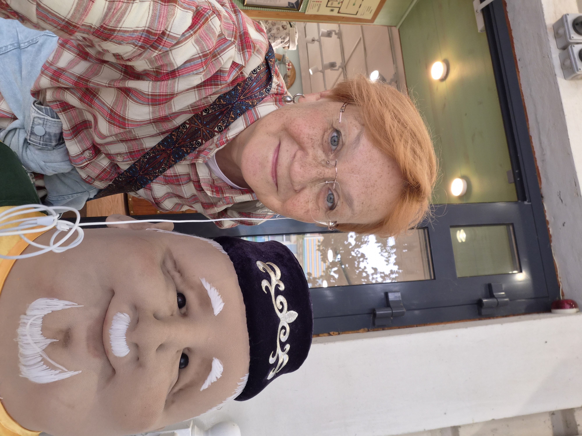 An outdoor, eye-level, medium shot shows a woman with short, ginger hair and glasses taking a selfie with a large, stylized doll. The woman is smiling and wearing a plaid shirt and light blue jeans. The doll has a round face with white eyebrows, a mustache, and a goatee. It is wearing a dark velvet cap with gold embroidery. In the background, there is a dark blue door with windows, and beyond that, a green-painted ceiling with lights. The scene appears to be in a marketplace or an outdoor stall.